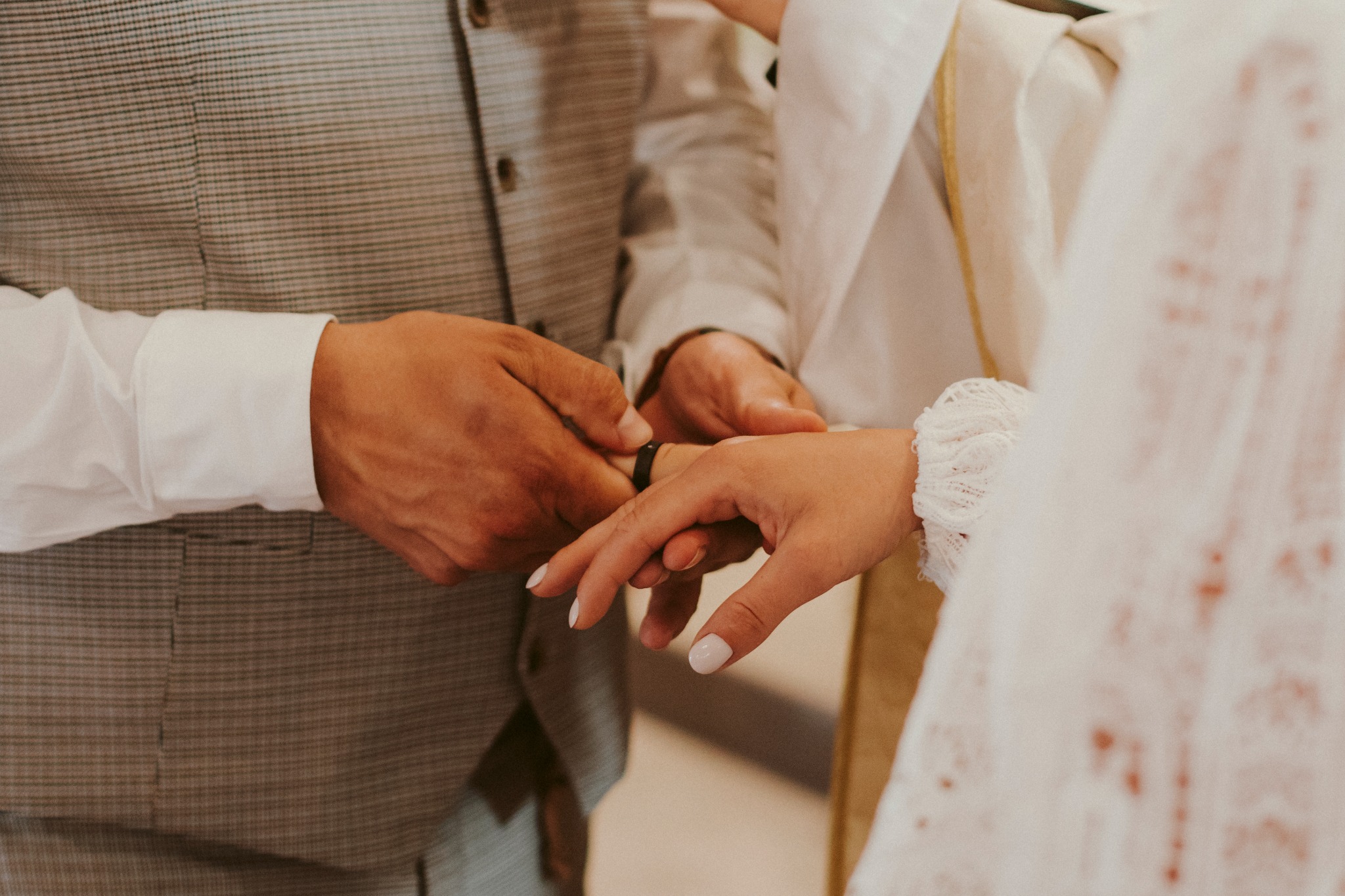 Zbliżenie dłoni podczas ceremonii ślubnej: mężczyzna zakłada czarną obrączkę na palec kobiety w białej, koronkowej sukni.