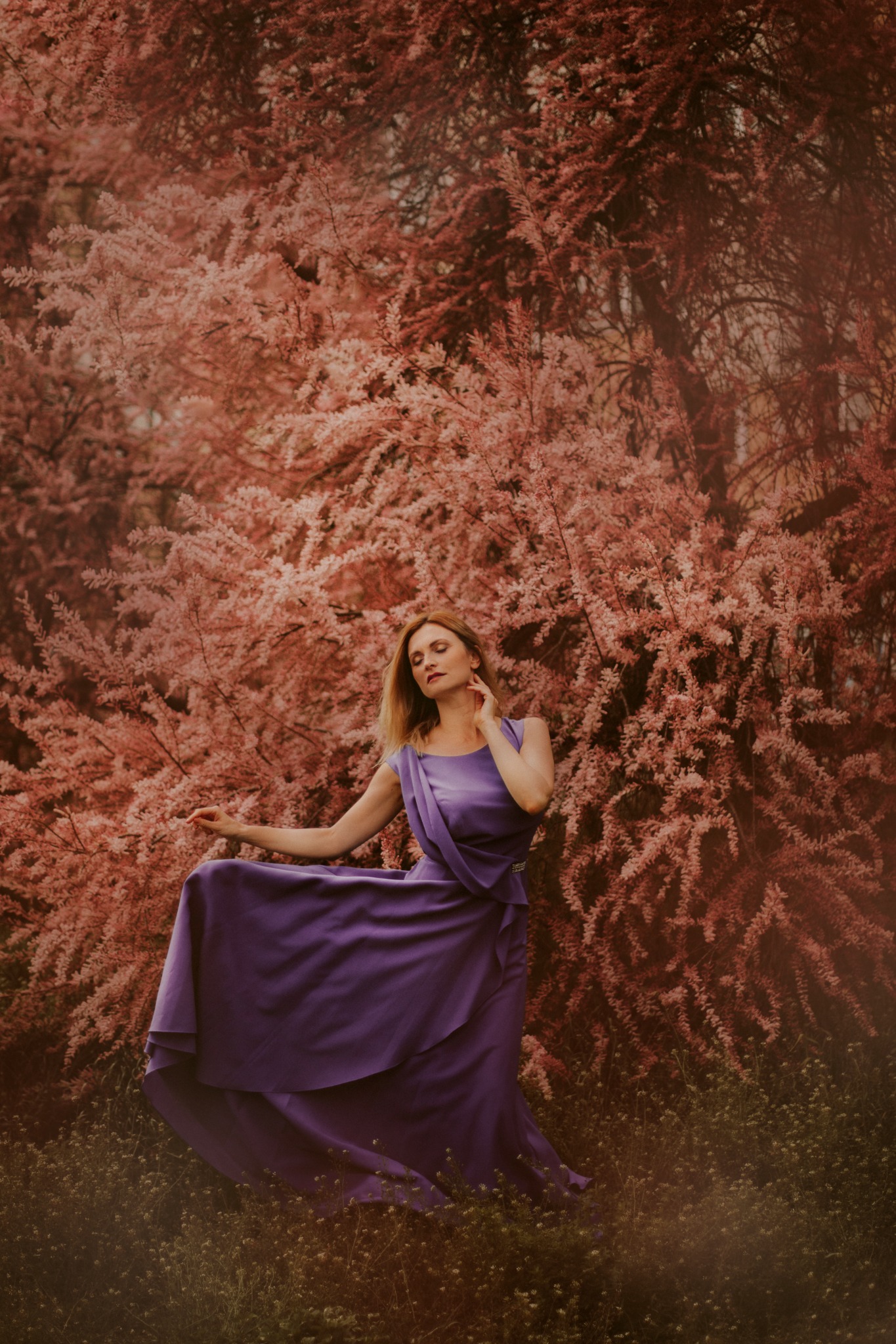 Kobieta w fioletowej sukni pozuje na tle kwitnącego drzewa o różowych kwiatach, delikatne światło podkreśla jej urodę.