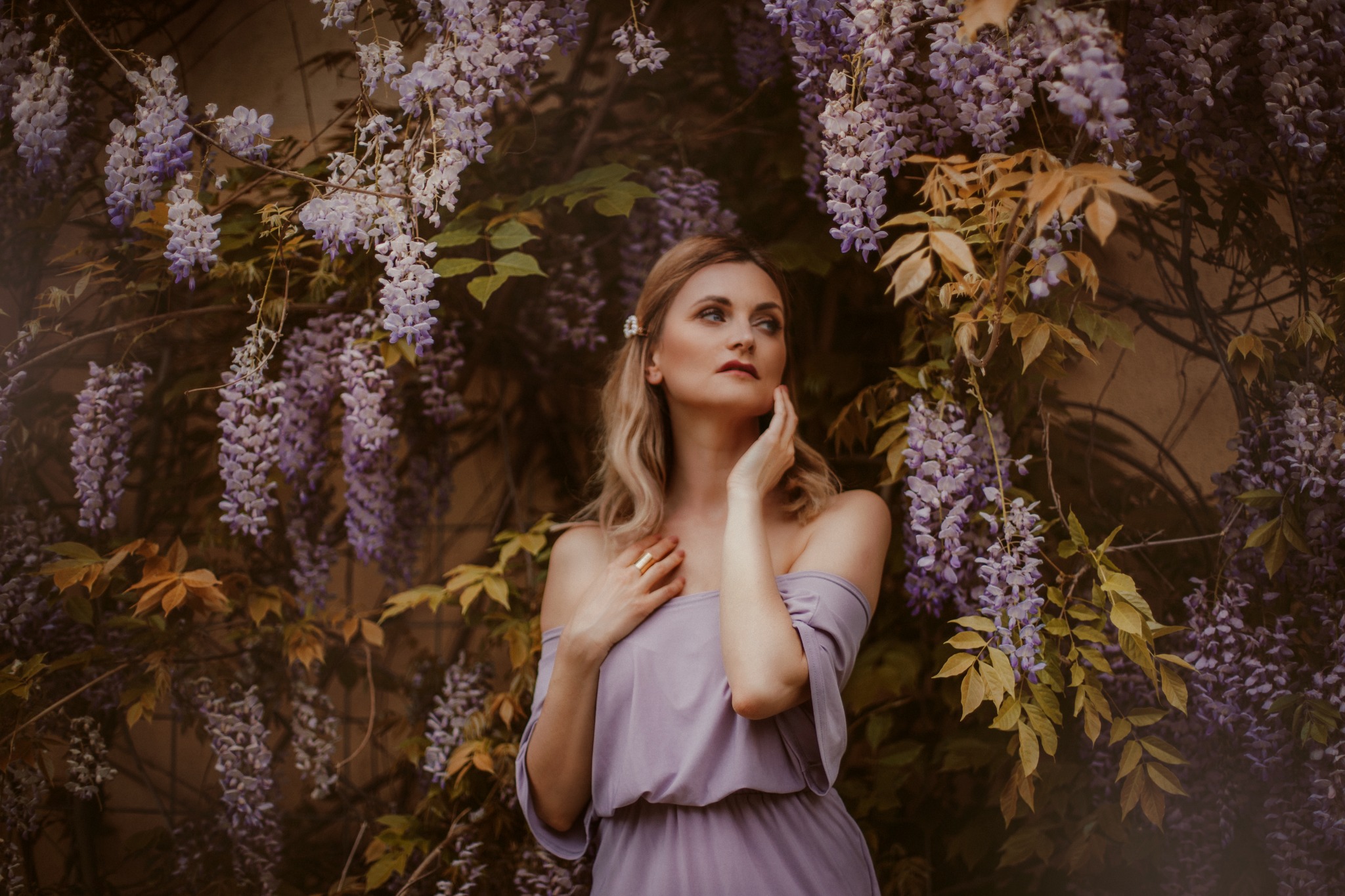 Kobieta w liliowej sukience pozuje na tle kwitnącej glicynii w Warszawie, z delikatnym światłem podkreślającym jej profil.