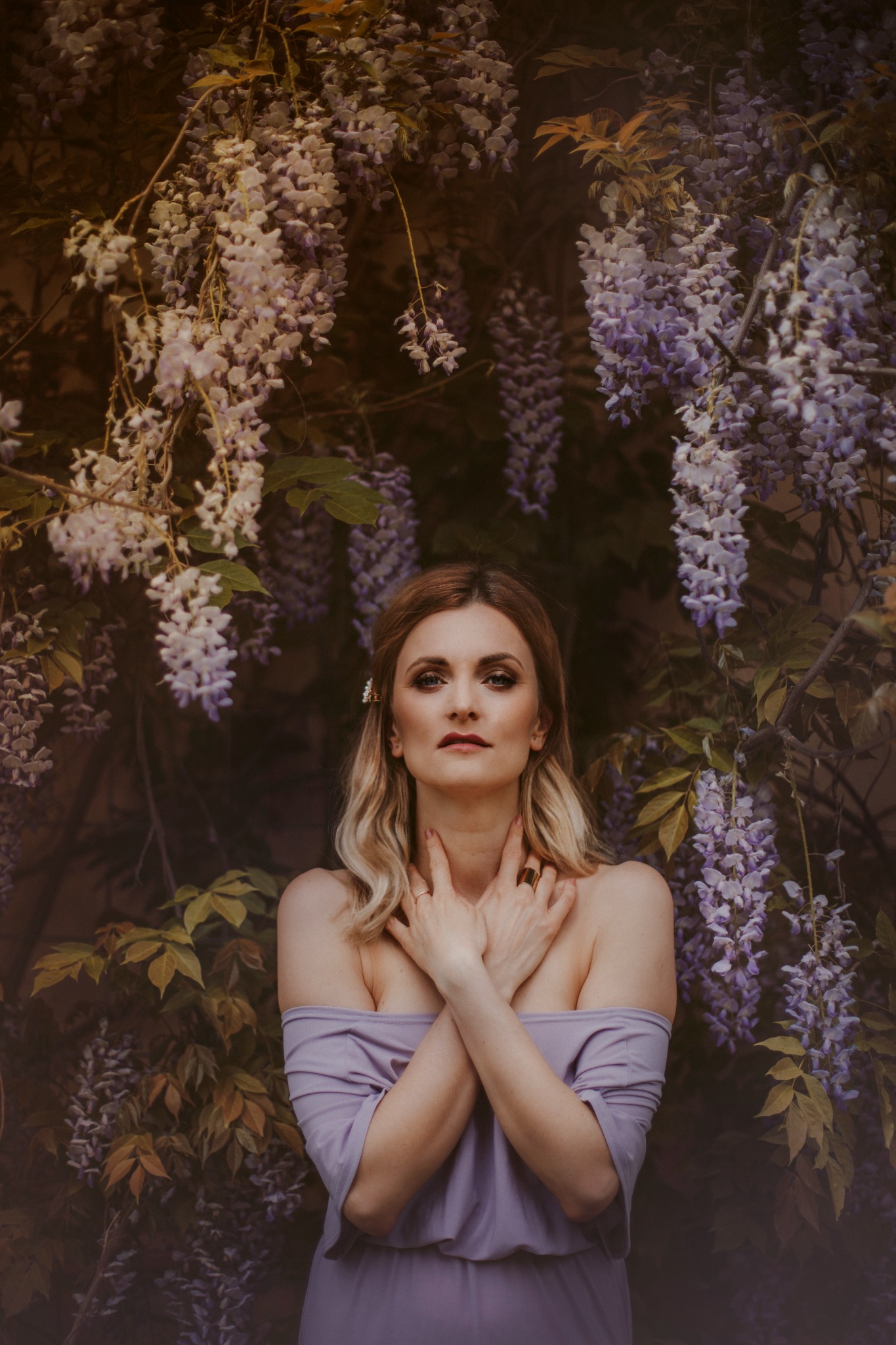 Portret kobiety w liliowej sukience na tle kwitnącej glicynii, ręce skrzyżowane na szyi, delikatny makijaż i upięcie włosów.