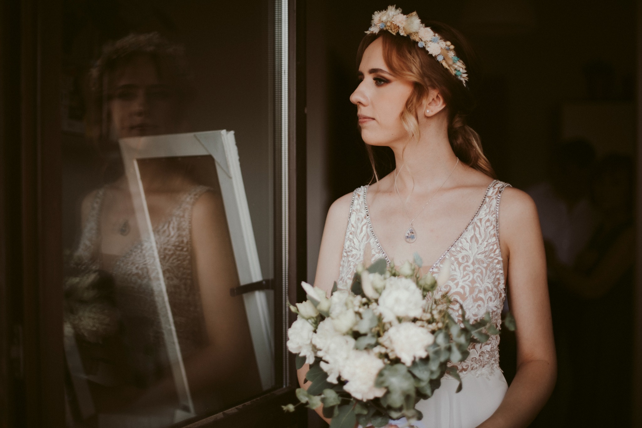 Panna młoda w koronie z kwiatów, z bukietem w rękach, spogląda w bok, a w szybie odbija się jej postać trzymająca białą ramkę.