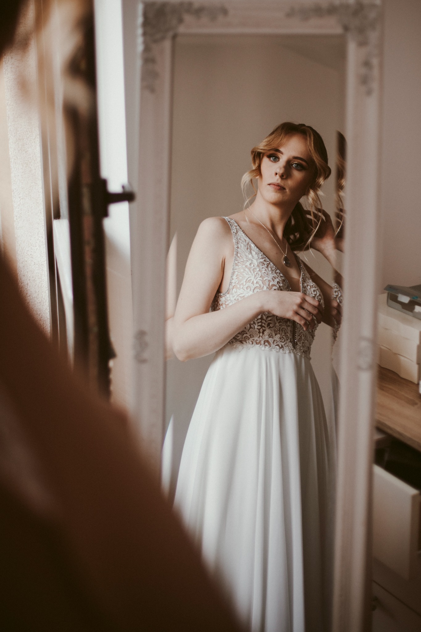 Kobieta w białej sukni z koronkową górą, poprawia włosy, widoczna w lustrze z ozdobną ramą.