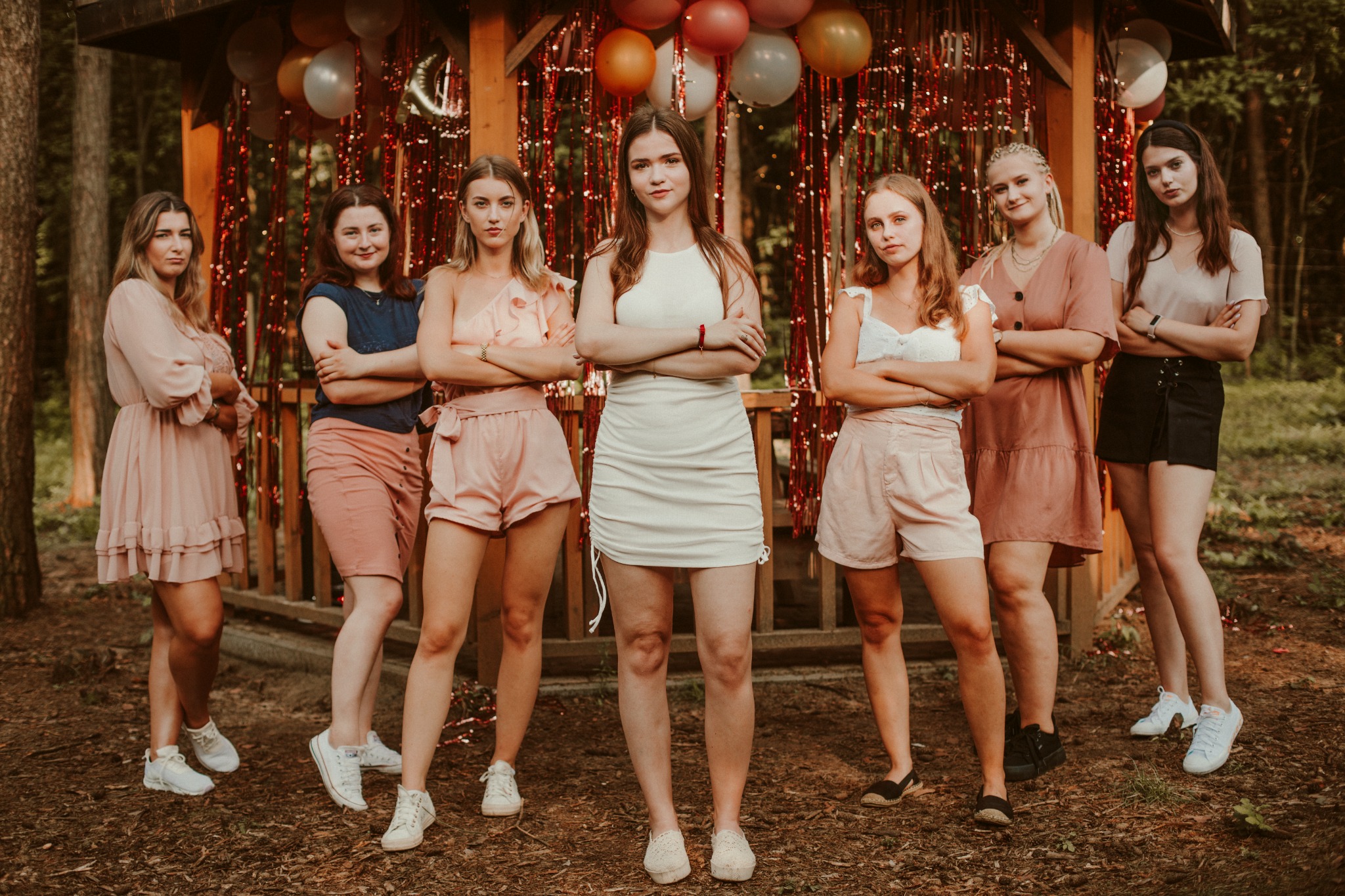 Grupa siedmiu młodych kobiet pozuje na zewnątrz, na tle drewnianej altany udekorowanej balonami i błyszczącymi girlandami, wszystkie z założonymi rękami, w różnorodnych letnich strojach i obuwiu.