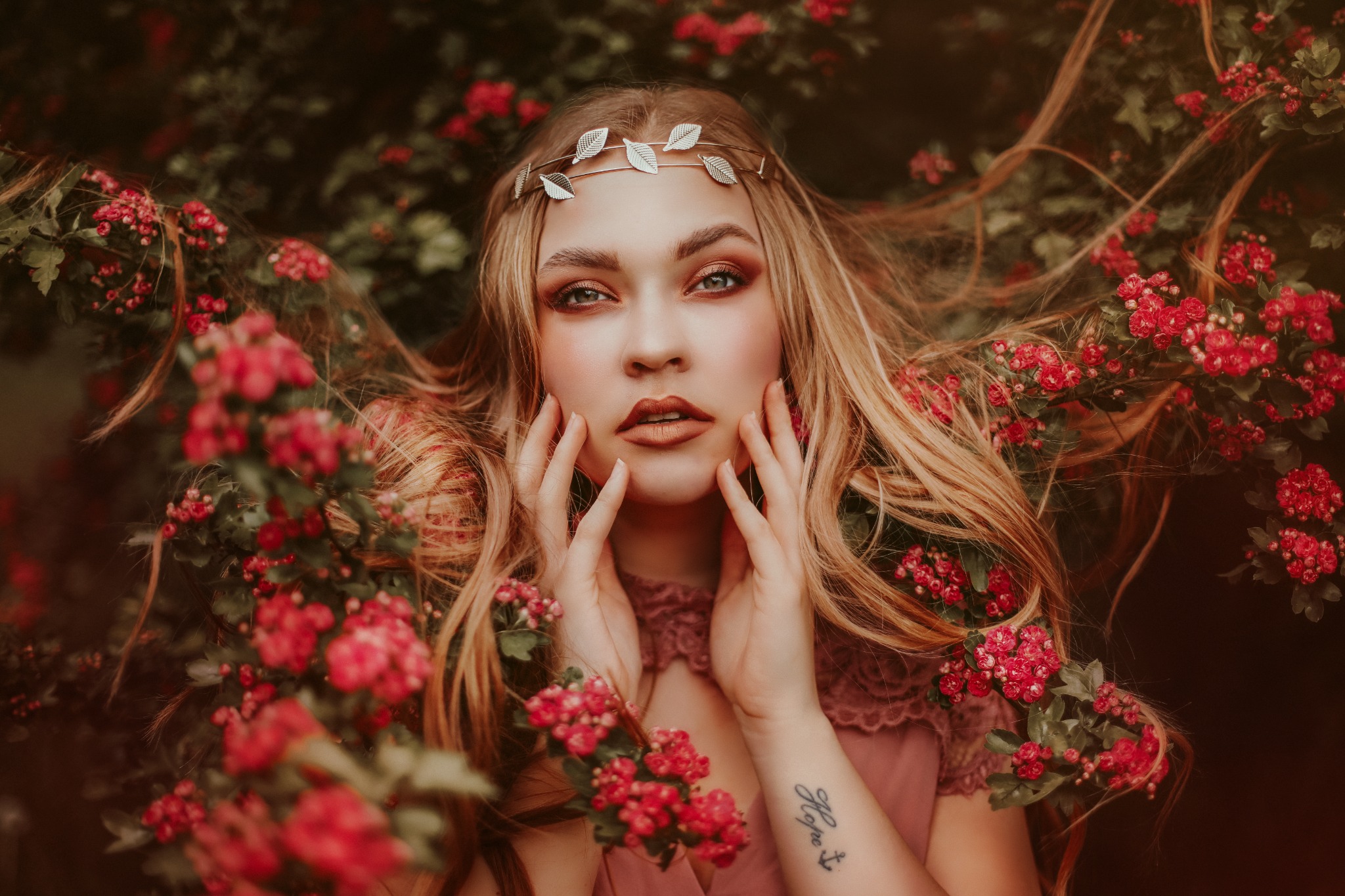 Portret kobiety z blond włosami, w sukience w kolorze pudrowego różu i z ozdobą z liści na głowie, otoczonej kwitnącymi gałązkami.