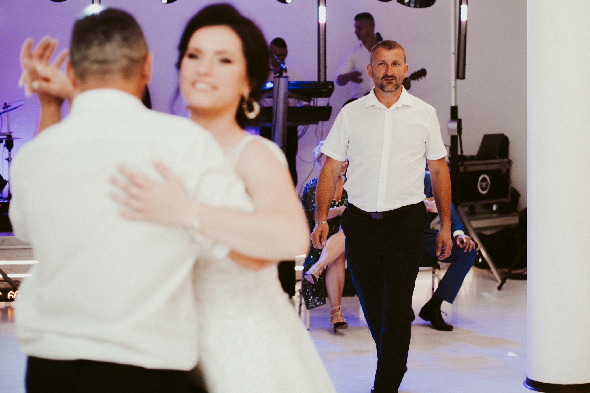Para młoda tańczy na parkiecie, w tle zespół muzyczny i goście weselni, mężczyzna w białej koszuli idzie w kierunku obiektywu.