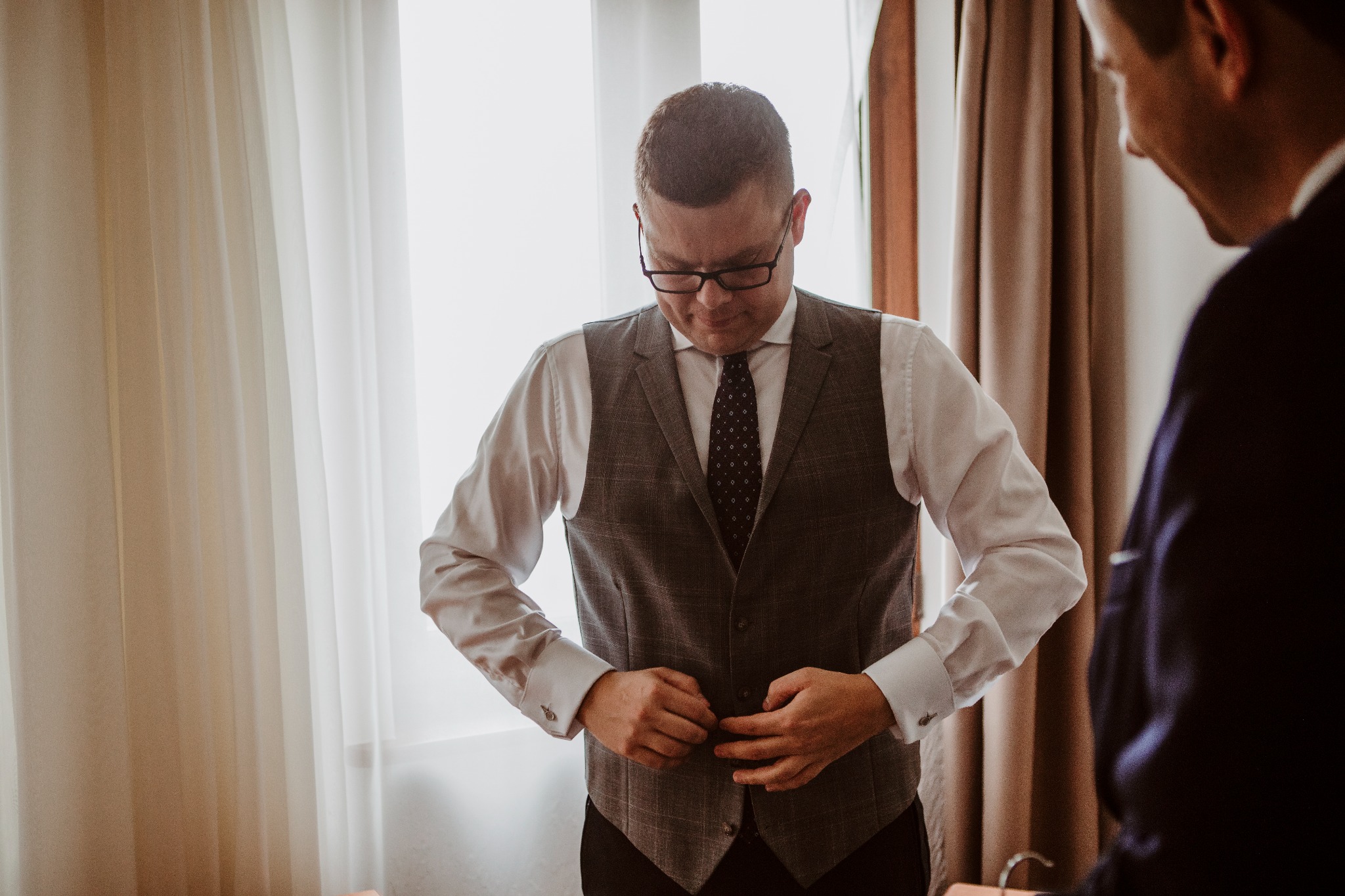 Mężczyzna w okularach zapina elegancką, kraciastą kamizelkę do garnituru, obok stoi druga osoba w garniturze, w tle okno z jasnymi zasłonami.