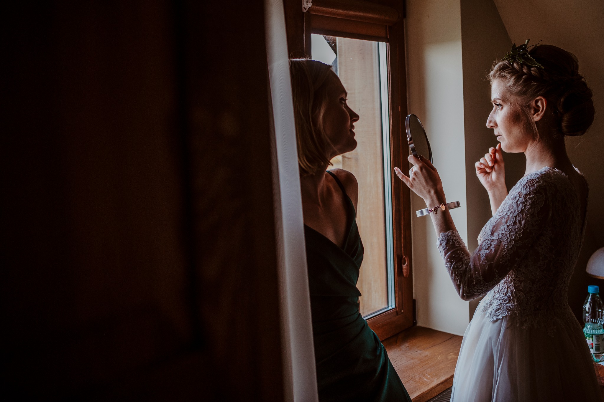 Panna młoda w koronkowej sukni, z wiankiem na głowie, przegląda się w lusterku trzymanym w dłoni, obok stoi druhna w zielonej sukni, obie przy oknie.