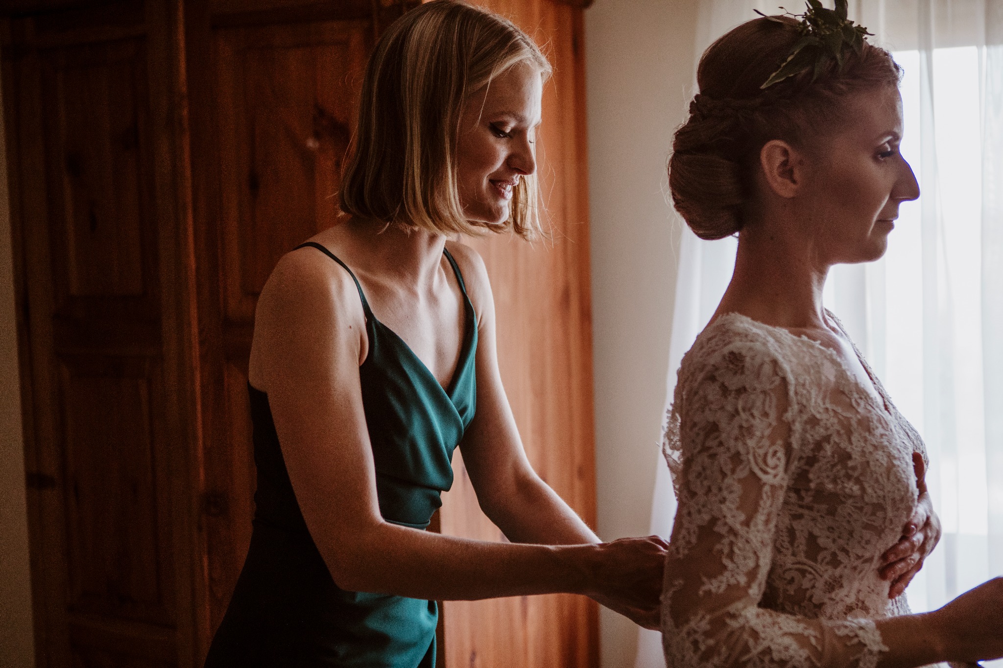 Panna młoda w koronkowej sukni ślubnej, poprawiana przez kobietę w szmaragdowej sukience na ramiączkach przed drewnianą szafą i oknem z białymi zasłonami.