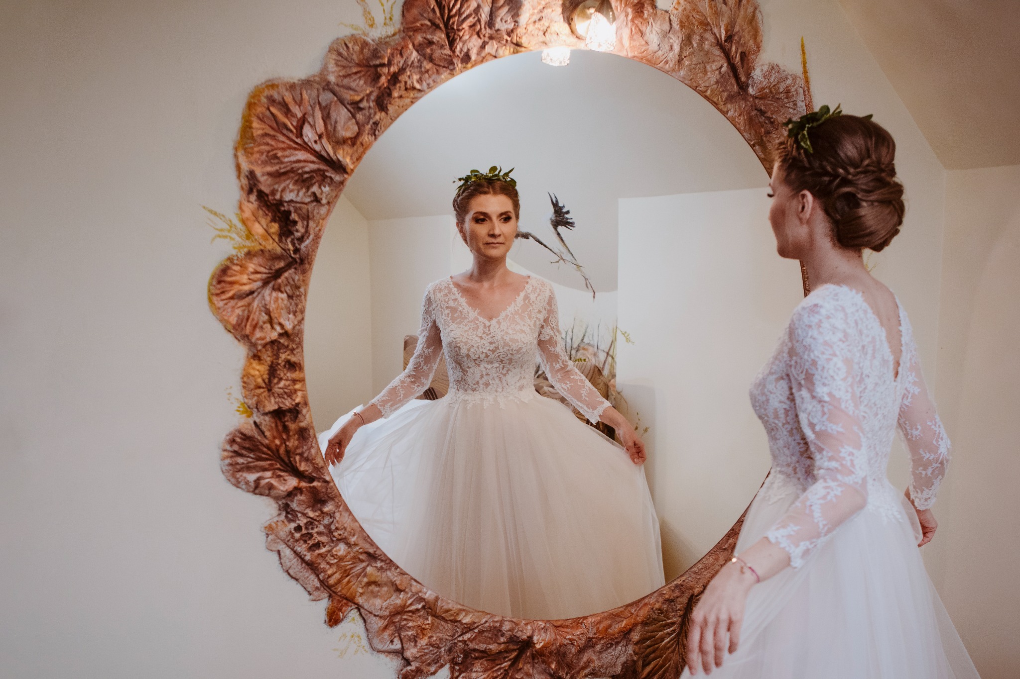 Panna młoda w koronkowej sukni ślubnej, z wiankiem na głowie, przegląda się w ozdobnym lustrze z motywem liści.