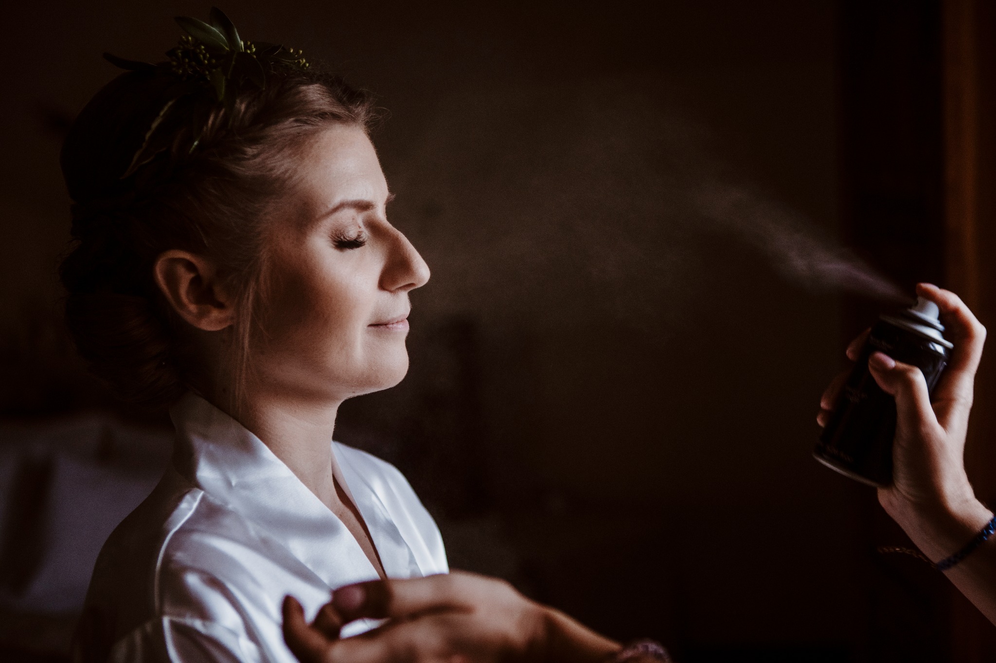 Kobieta w białym szlafroku z zamkniętymi oczami, podczas aplikacji sprayu do włosów, z delikatną ozdobą z zielonych liści we włosach.