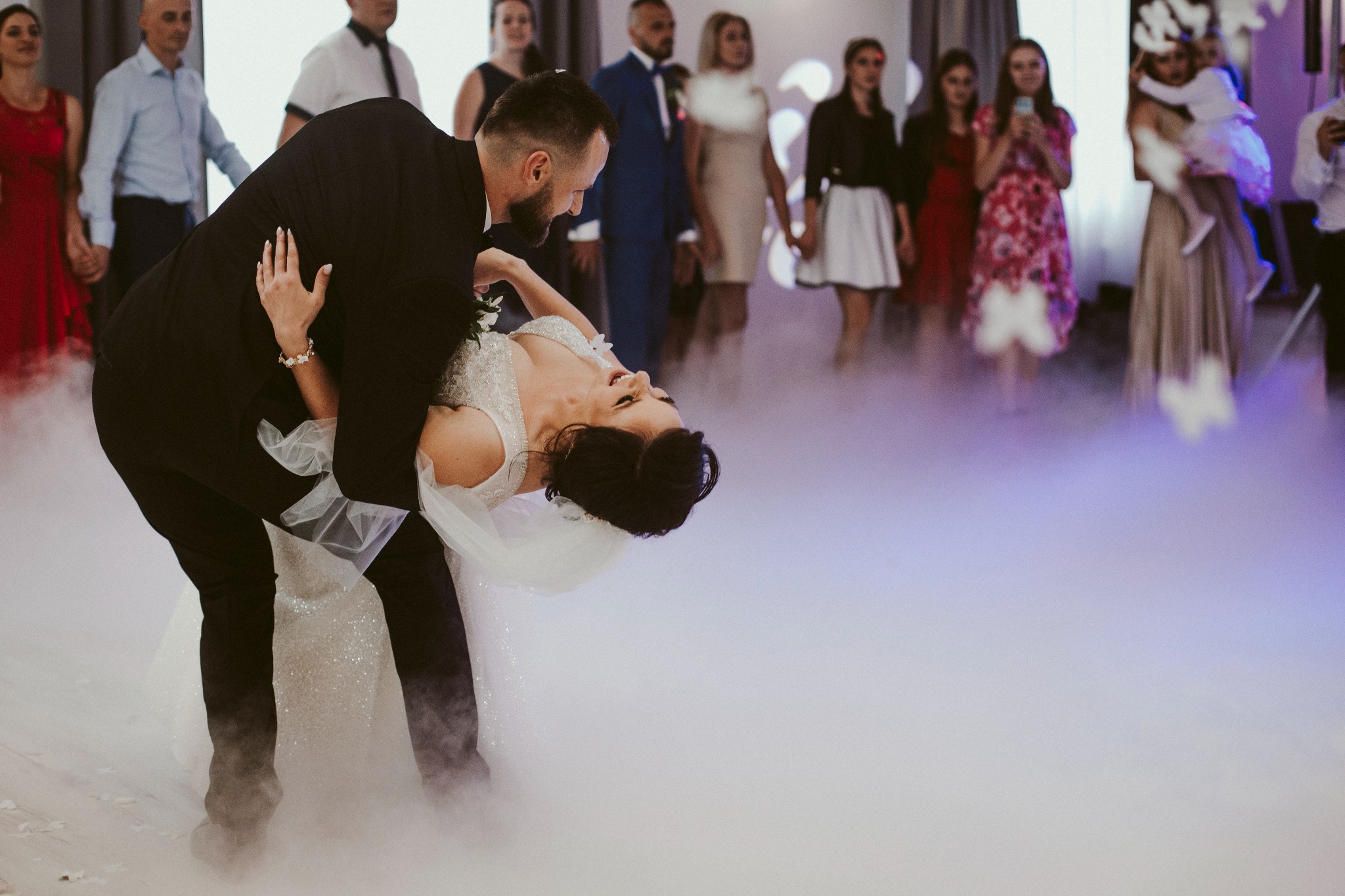 Para młoda w tańcu, panna młoda w białej sukni z tiulowym welonem odchylona do tyłu, pan młody w czarnym garniturze podtrzymuje ją, w tle goście weselni i ciężki dym na parkiecie.