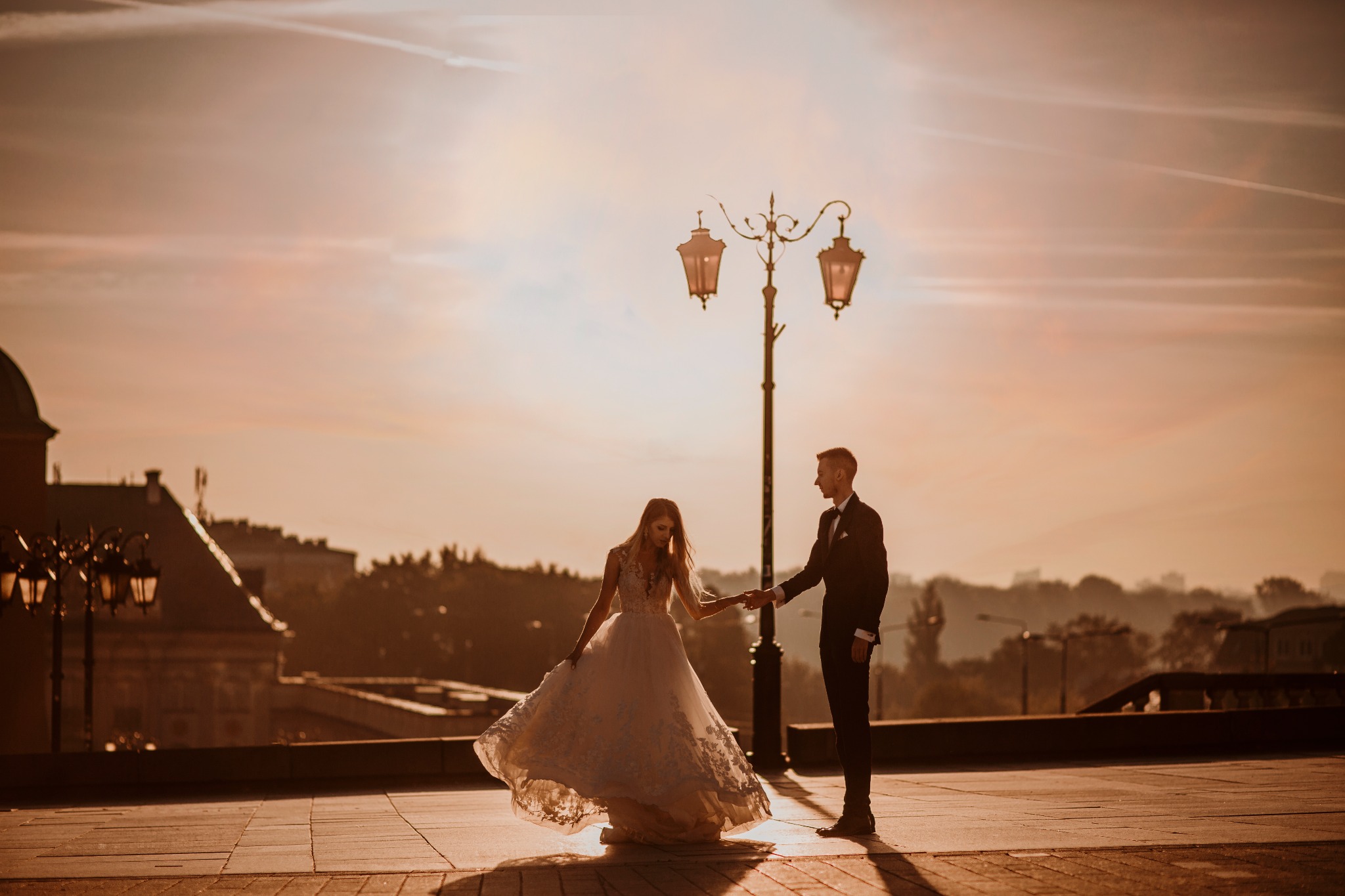 Para młoda trzymająca się za ręce na tle zachodzącego słońca i miejskiej architektury, oświetlona latarnią