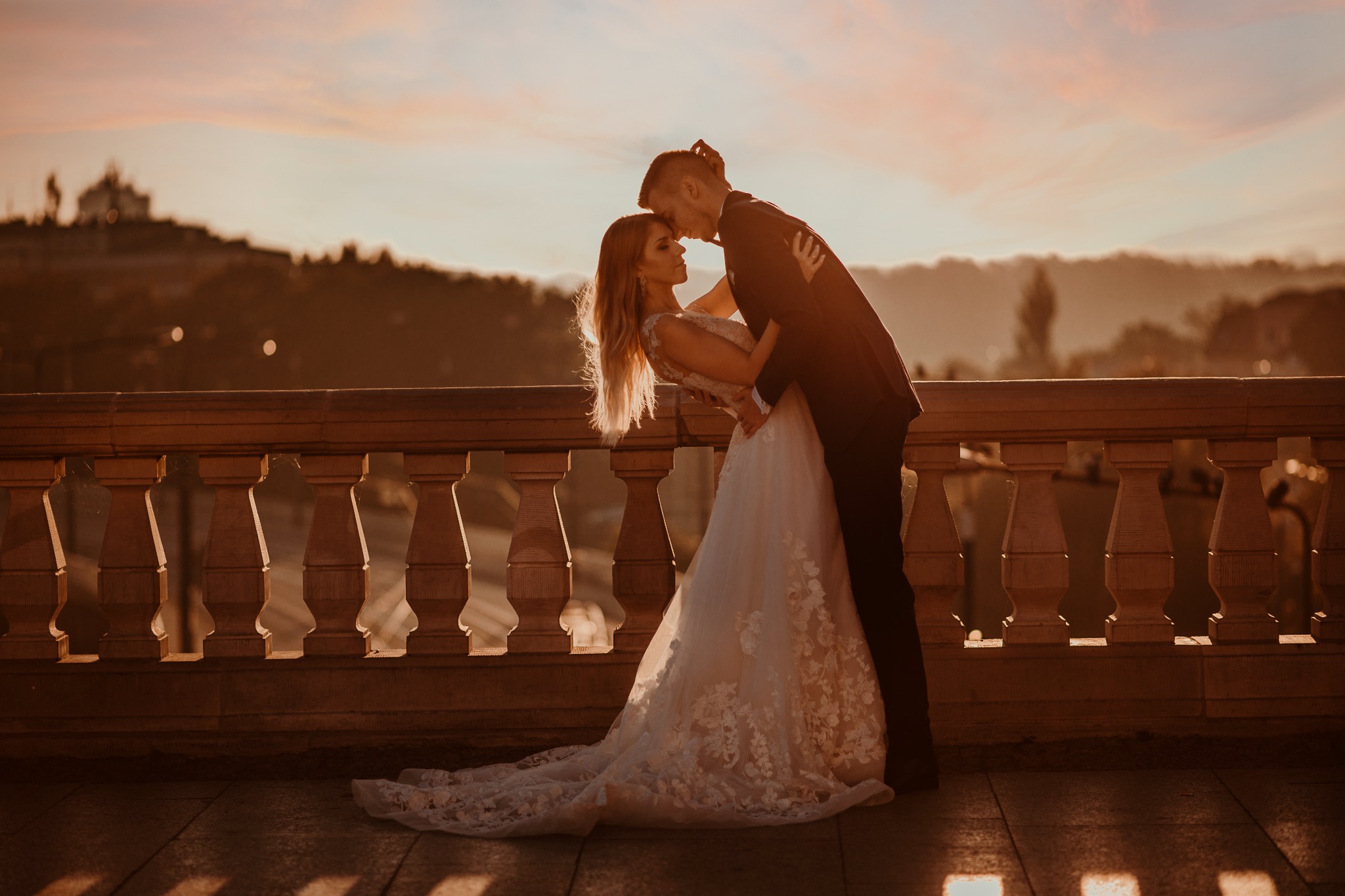 Para młoda w objęciach na tle zachodzącego słońca i panoramy miasta, panna młoda w białej sukni z koronkowymi aplikacjami, pan młody w ciemnym garniturze.