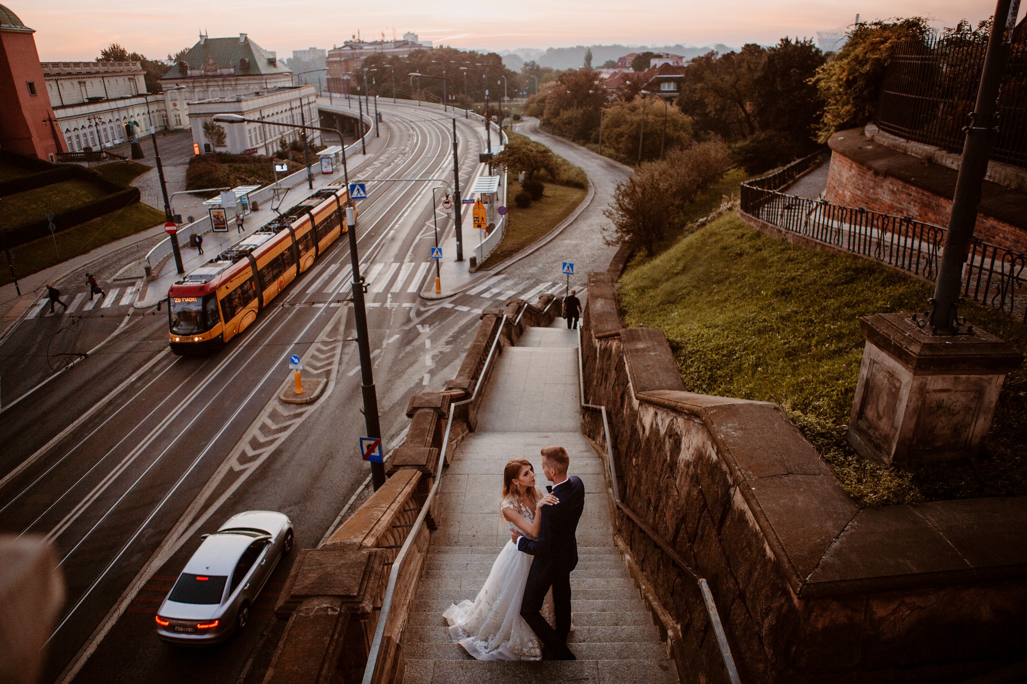 Para młoda obejmuje się na schodach z widokiem na miasto, tramwaj i samochód w tle, podczas sesji zdjęciowej w Warszawie.