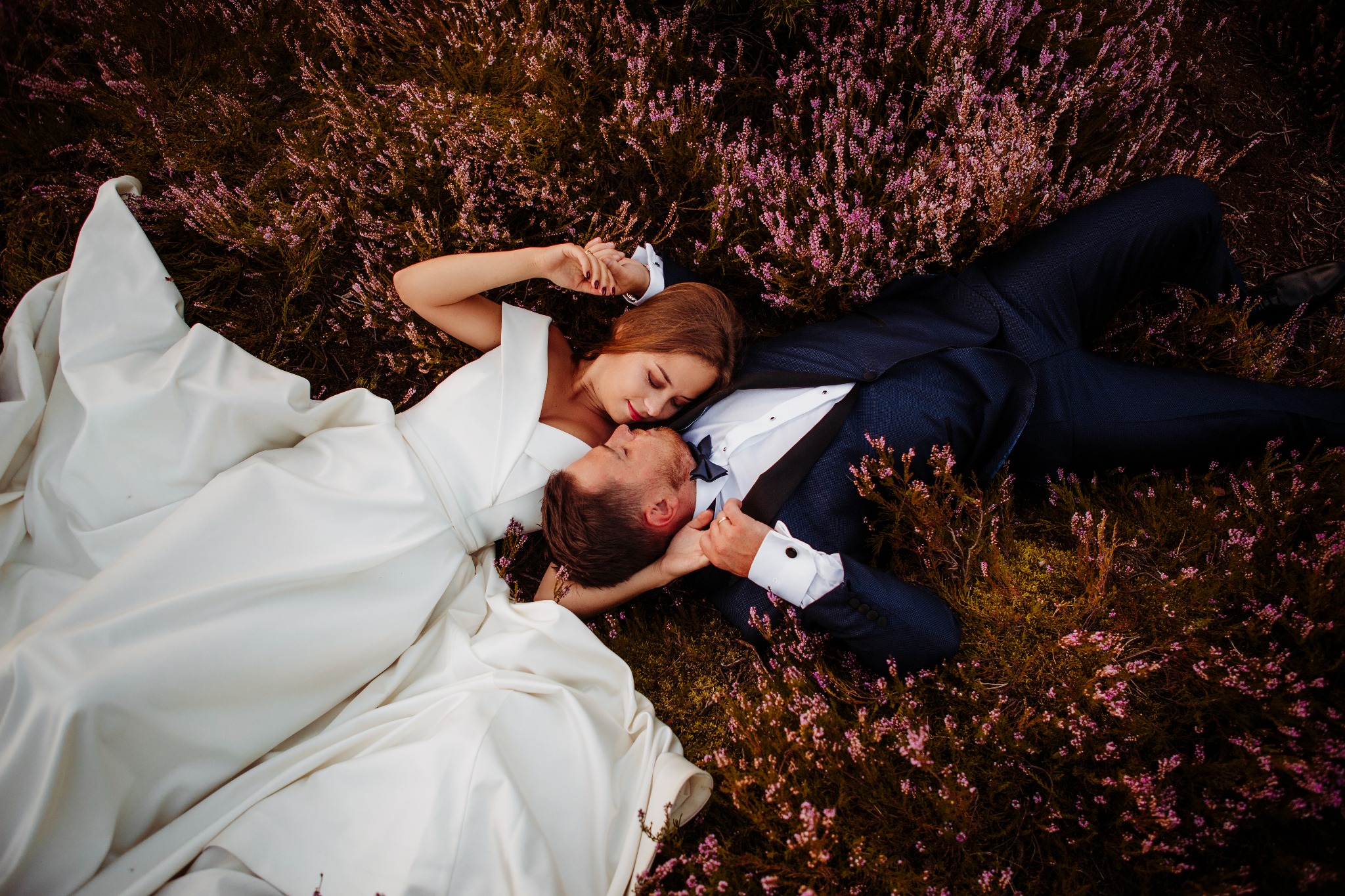 Zakochana para, panna młoda w białej sukni i pan młody w granatowym garniturze, leżą na wrzosowisku, romantyczny kadr z góry.