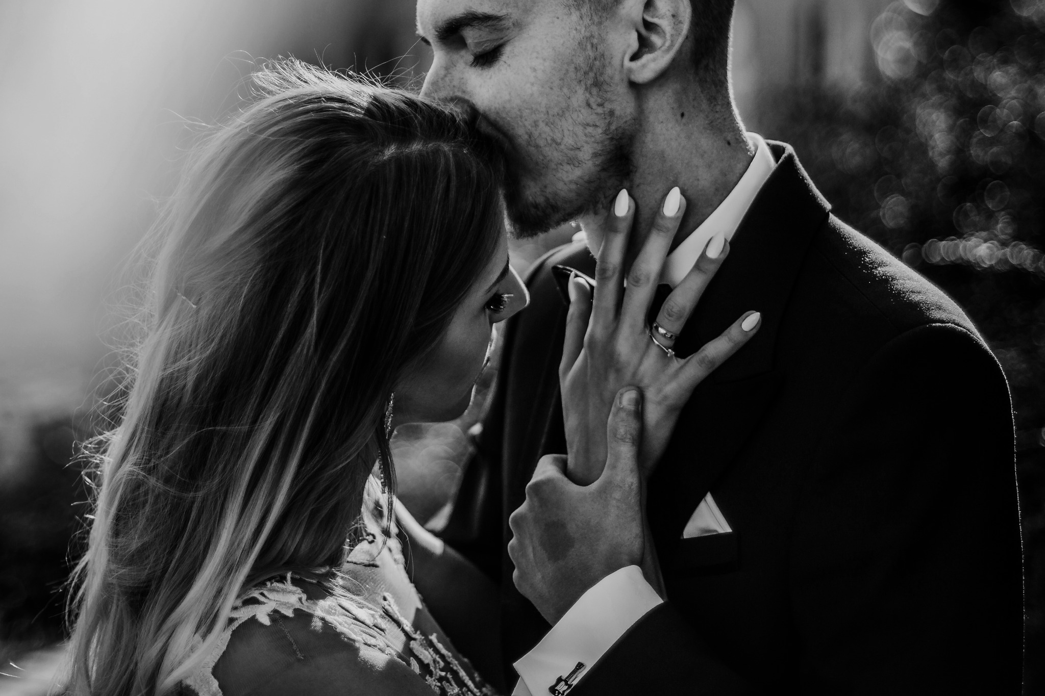 Czarno-białe zbliżenie: mężczyzna w garniturze całuje kobietę z długimi włosami w czoło, pierścionek zaręczynowy na palcu kobiety, mankiety z motywem gitary.