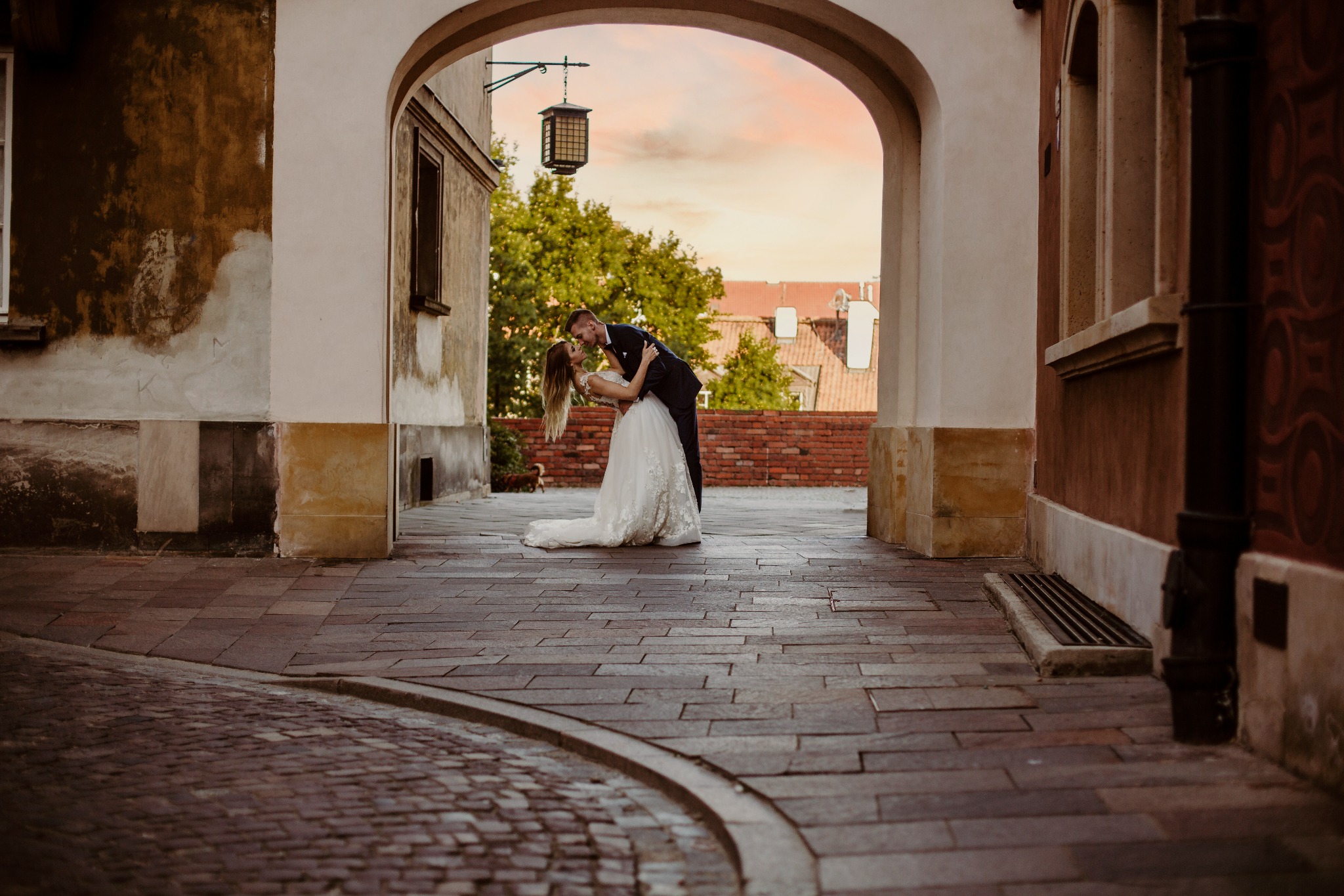 Para młoda w objęciach pod kamiennym łukiem, panna młoda w białej sukni z koronkowymi aplikacjami, pan młody w ciemnym garniturze, w tle błękitne niebo o zachodzie słońca i staromiejska zabudowa.