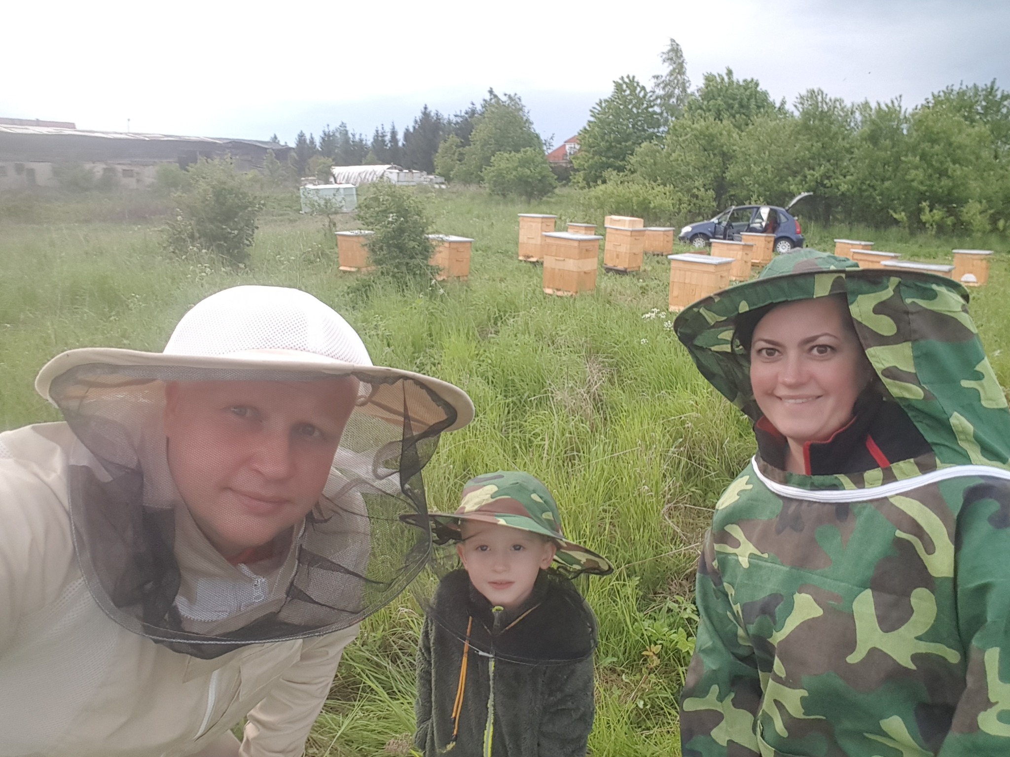 Rodzina pszczelarzy w strojach ochronnych na tle uli w plenerze, z samochodem w tle