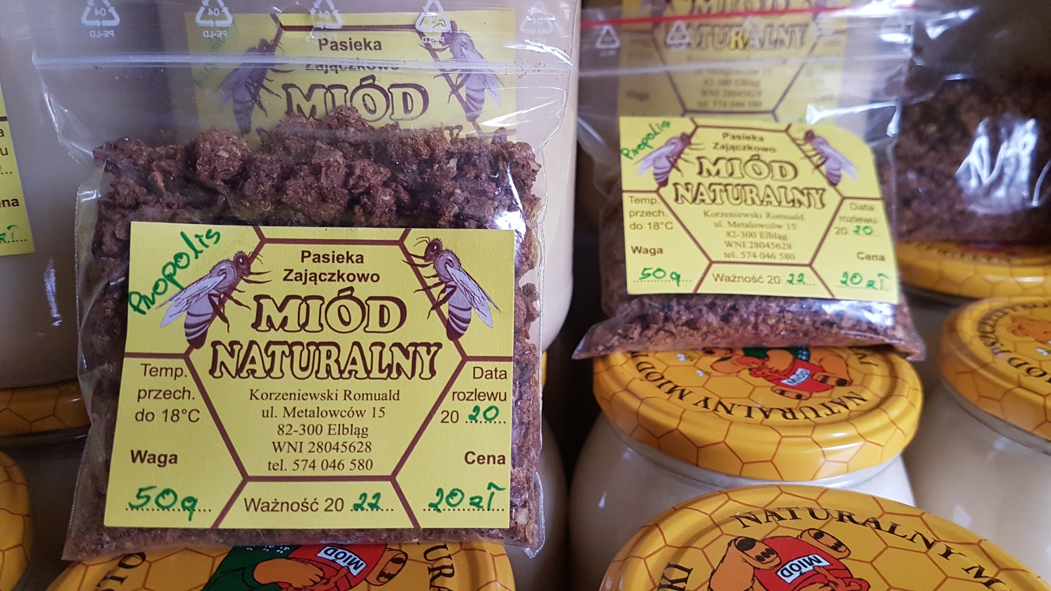 Paczki z pyłkiem pszczelim z propolisem, etykieta z nazwą pasieki i informacjami o produkcie, widoczne słoiki z miodem w tle.