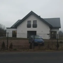 Nowoczesny dom jednorodzinny z szarą blachodachówką, białymi ścianami i ciemnymi oknami, częściowo zasłonięty przez niebieski samochód i metalowe ogrodzenie w pochmurny dzień.