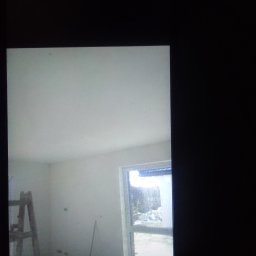 Anna Gębska - Pokój w trakcie prac wykończeniowych: białe ściany i sufit, drewniana drabina oparta o ścianę, widok na zewnątrz przez okno, ślady po szpachlowaniu widoczne na ścianie.