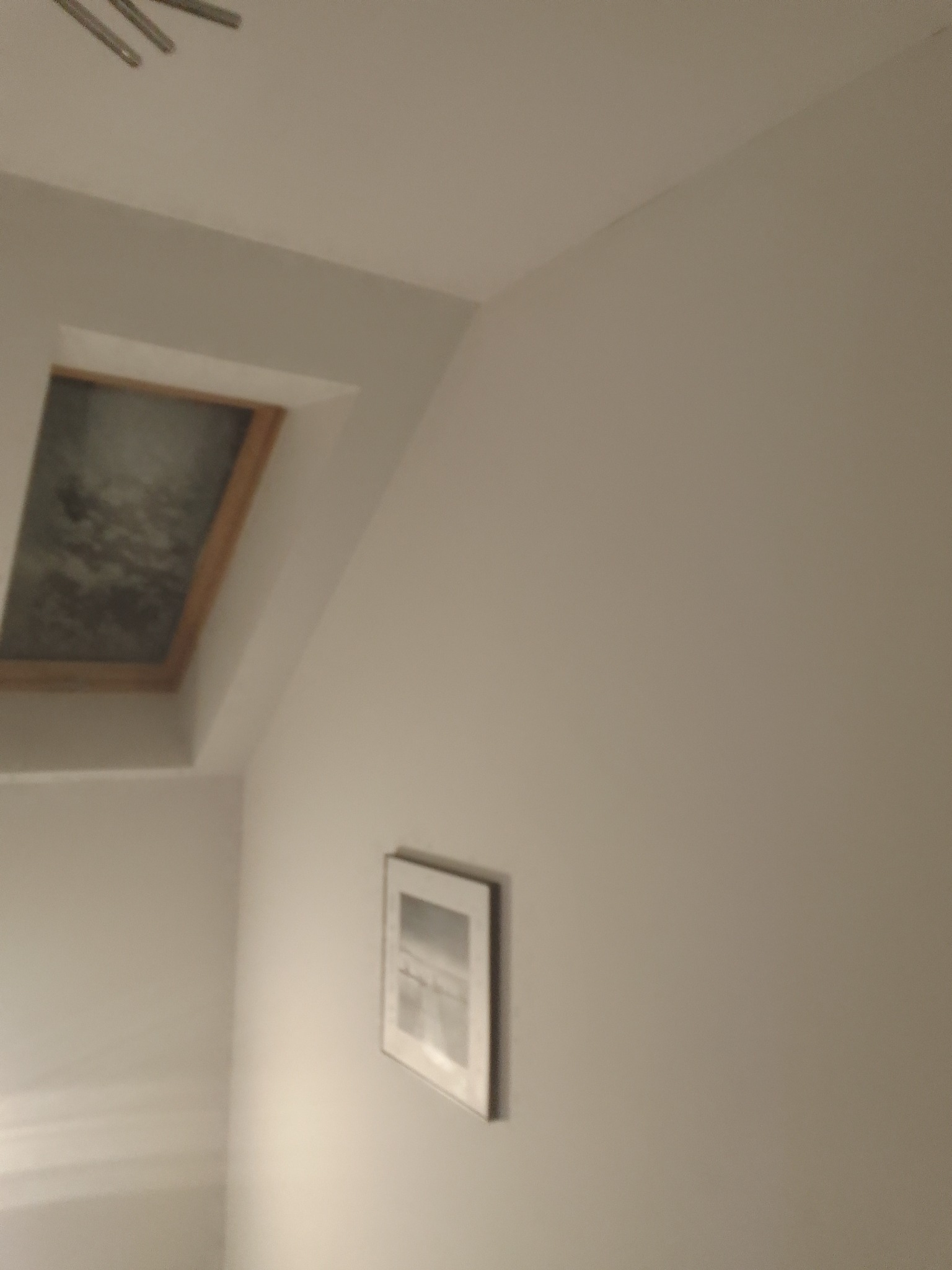 Poddasze z oknem dachowym i świeżo wykończoną, gładką ścianą w kolorze złamanej bieli, na której wisi obrazek w ramce.