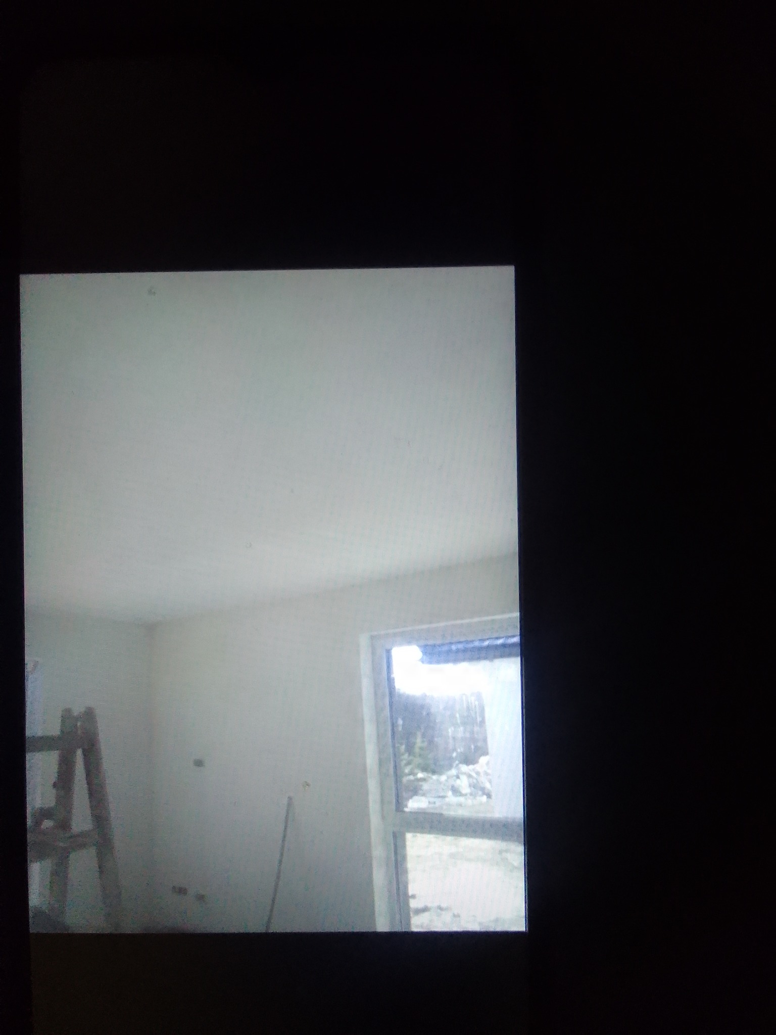 Pokój w trakcie prac wykończeniowych: białe ściany i sufit, drewniana drabina oparta o ścianę, widok na zewnątrz przez okno, ślady po szpachlowaniu widoczne na ścianie.