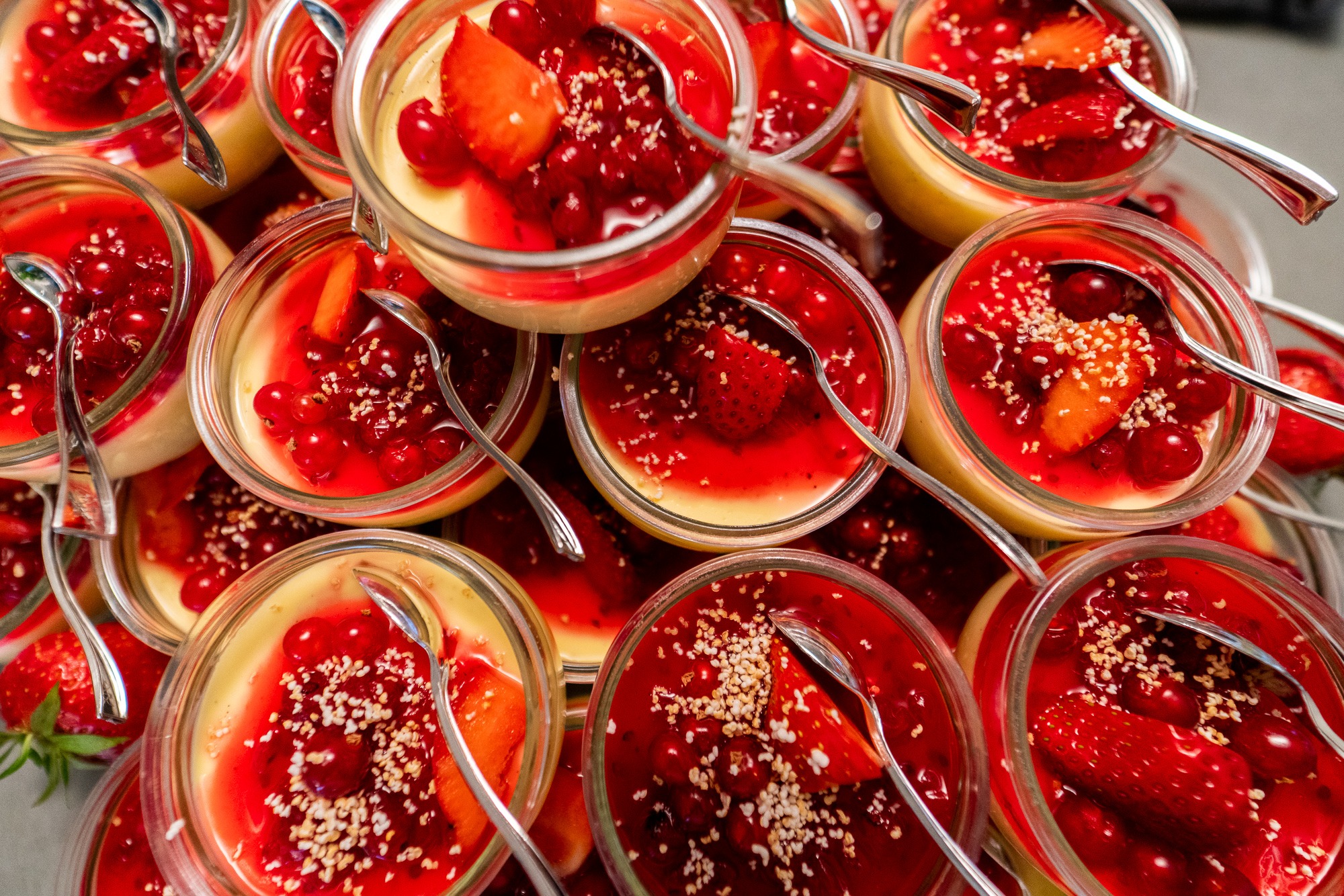 Deser panna cotta w szklanych słoiczkach, polany czerwoną galaretką z owocami leśnymi i posypany sezamem, każda porcja z małą srebrną łyżeczką.