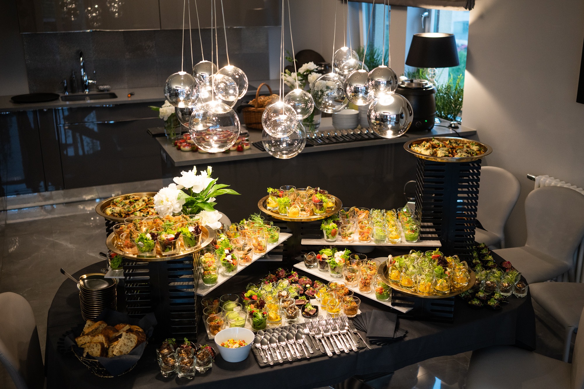 Elegancki stół cateringowy z różnorodnymi przekąskami w szklanych naczyniach, pizza na złotych paterach, białe kwiaty w wazonie i wiszące szklane lampy kuliste, nowoczesne wnętrze.