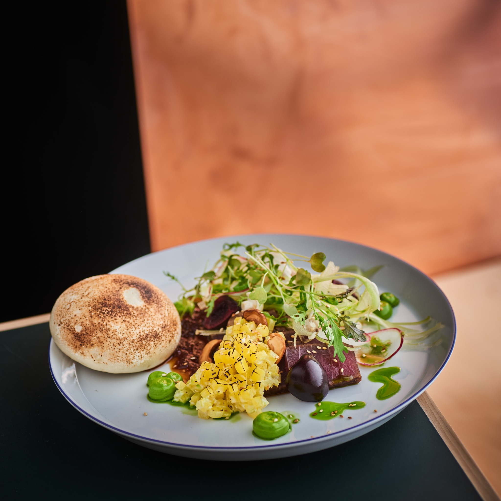 Elegancko podane danie z tuńczykiem, sałatką z rzodkiewką i sosem wasabi na białym talerzu z granatową obwódką, obok mała bułeczka z posypką, na tle czarnego i miedzianego tła.