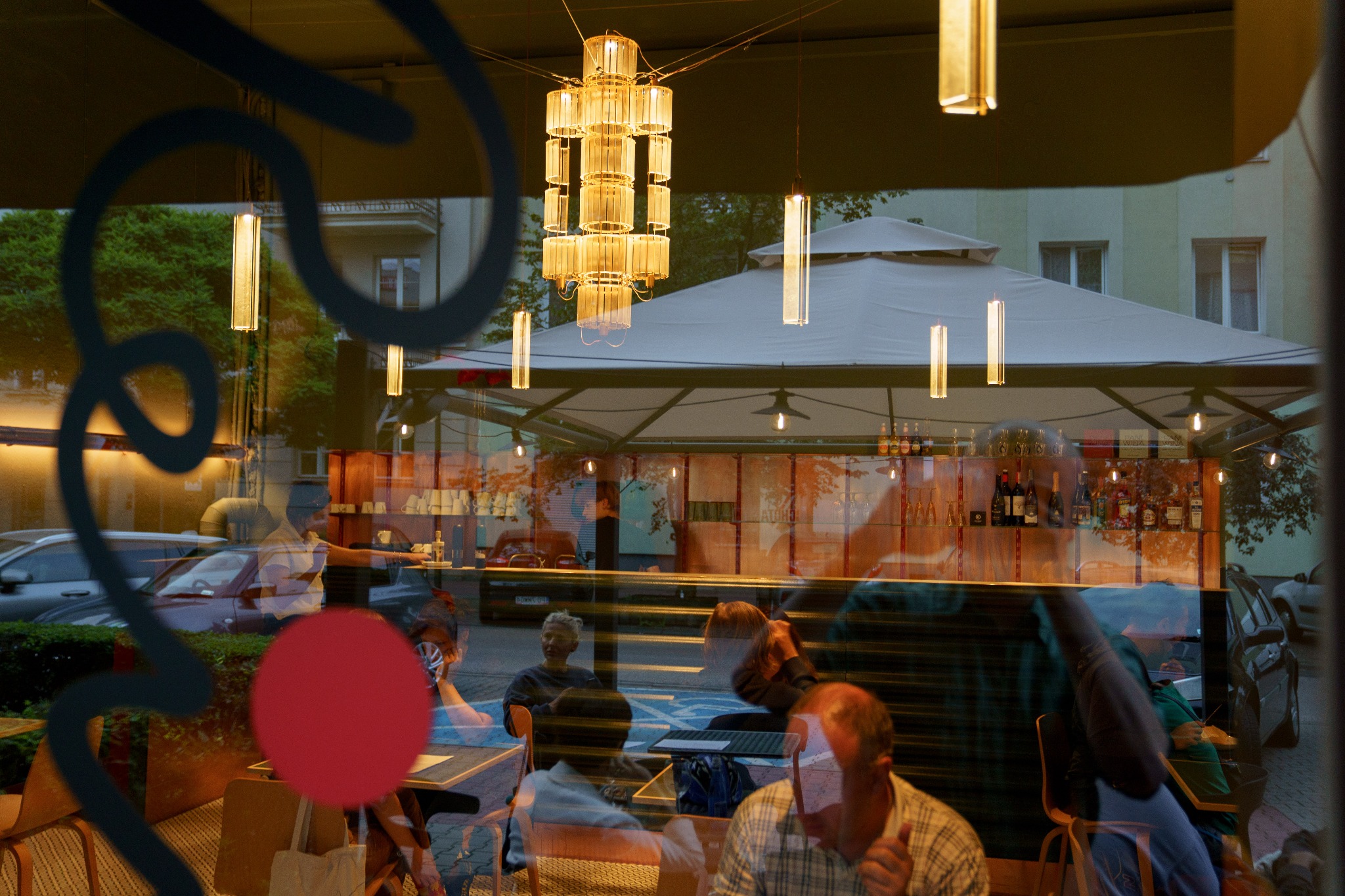 Widok przez witrynę restauracji na klientów siedzących przy stolikach, z odbiciem ulicy i dekoracyjnym oświetleniem wewnątrz i na zewnątrz.