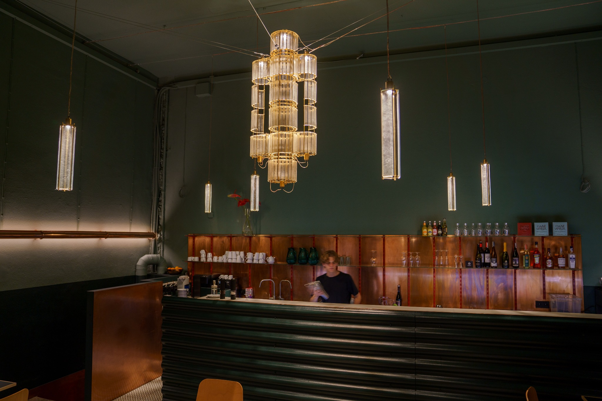 Wnętrze baru z zielonymi ścianami, miedzianym barem i półkami z napojami, oświetlone ozdobnymi lampami wiszącymi, za barem stoi osoba.