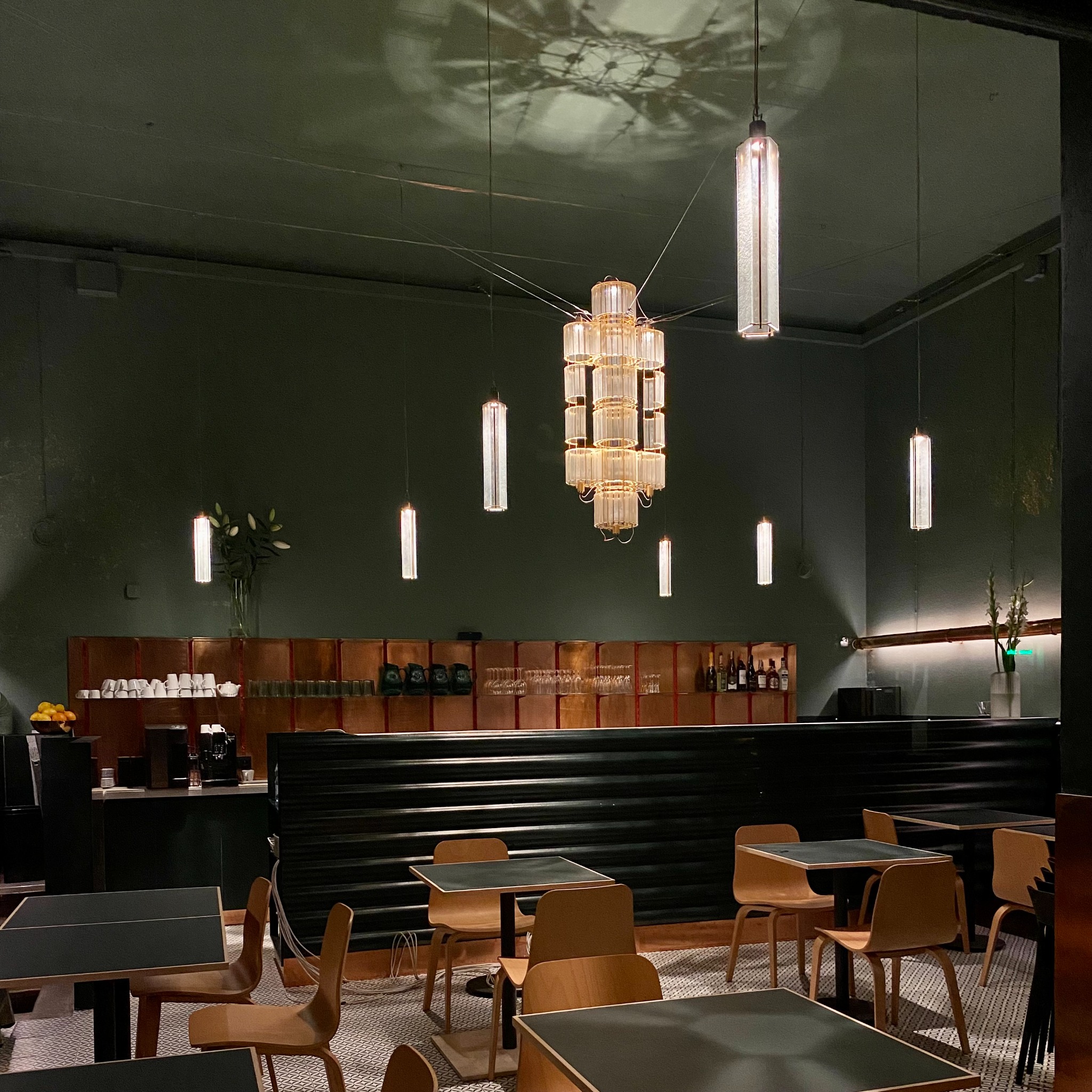 Wnętrze restauracji z ciemnozielonymi ścianami, drewnianymi krzesłami i stołami z czarnymi blatami, oświetlone ozdobnymi lampami wiszącymi i barem w tle z wyposażeniem.