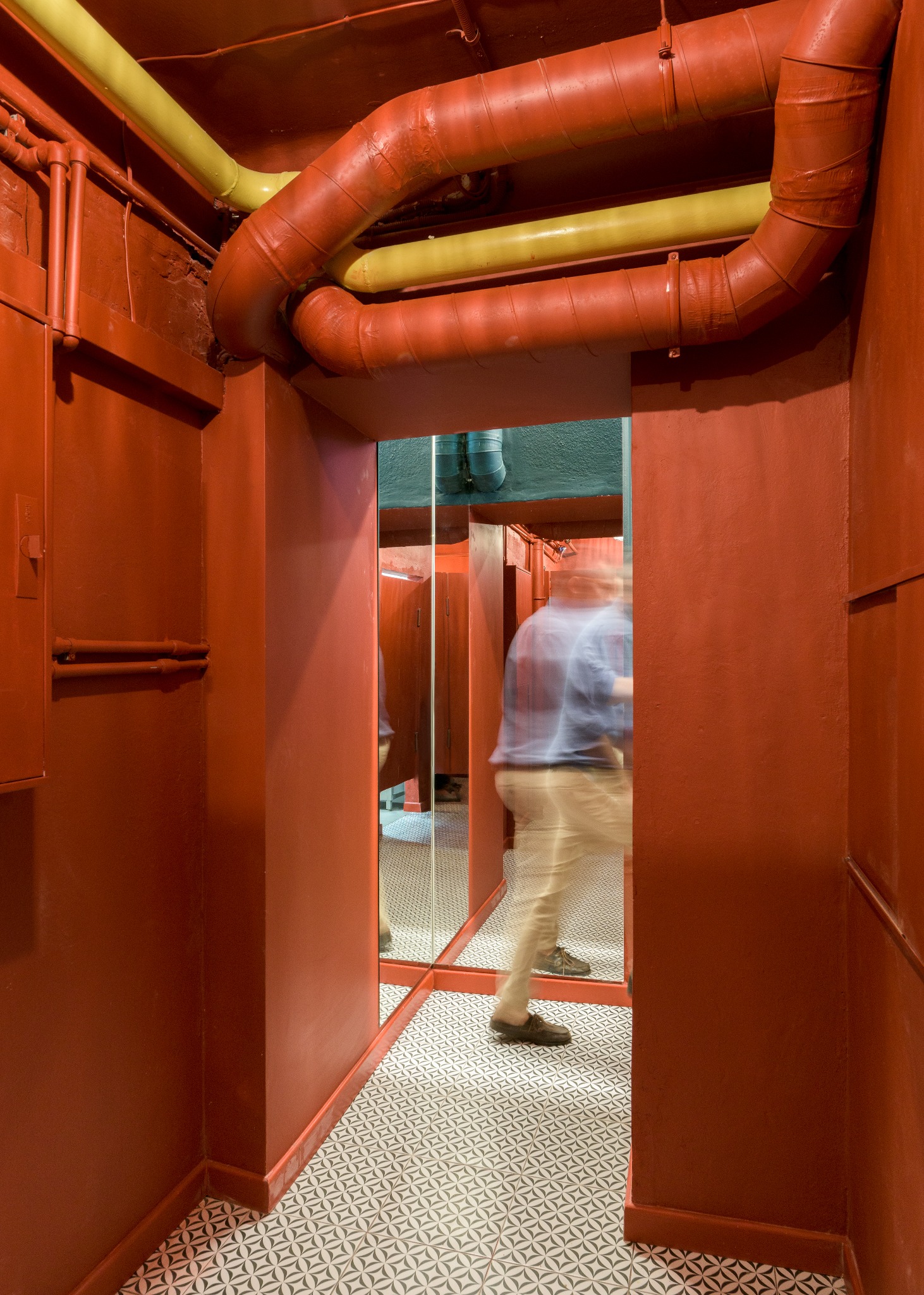 Pomieszczenie pomalowane na czerwono z widocznymi żółtymi i czerwonymi rurami pod sufitem, wzorzysta podłoga, lustro odbijające kabiny i zamazana sylwetka osoby w ruchu w tle.
