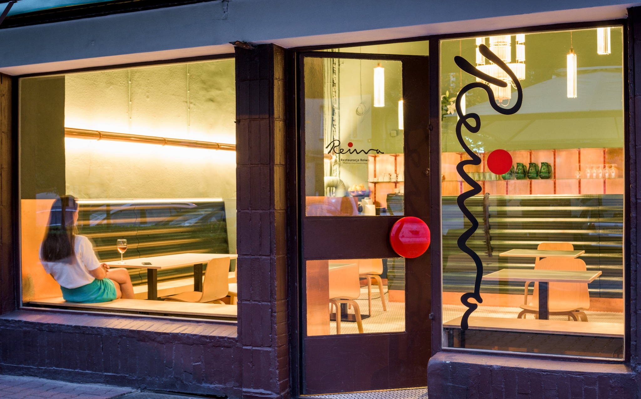 Widok z zewnątrz na restaurację z osobą siedzącą przy stoliku w oknie, przez szybę widoczne wnętrze z elementami dekoracyjnymi i stolikami.