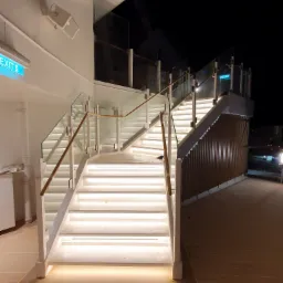 Nowoczesne schody z podświetlanymi stopniami i szklanymi balustradami z drewnianymi poręczami na statku nocą.