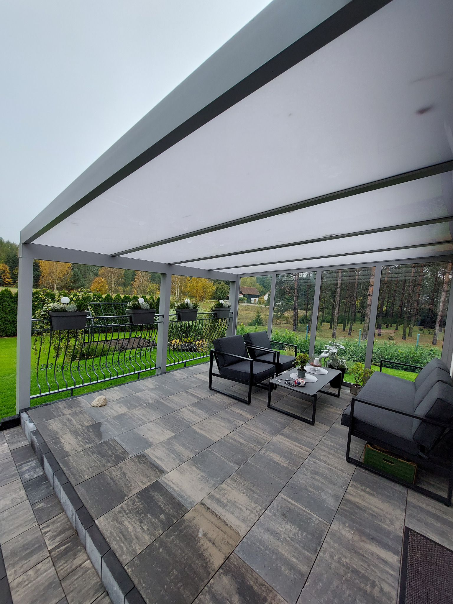 Zadaszony taras z aluminiową konstrukcją, boczną ścianą ze szkła ESG VSG i dachem z poliwęglanu litego 8mm, wyposażony w meble ogrodowe na tle zieleni.