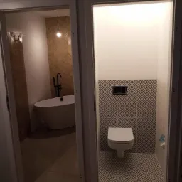 Dwa pomieszczenia łazienkowe widoczne przez otwarte drzwi: po lewej wanna wolnostojąca i ściana z mozaiką w odcieniach beżu, po prawej toaleta podwieszana z geometryczną mozaiką na ścianie i podłodze.