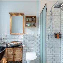 Nowoczesna łazienka z czarną wanną, umywalką z kamienia na drewnianej szafce, białą toaletą, kabiną prysznicową i drabinką na ręczniki, wykończona białymi płytkami imitującymi cegłę.