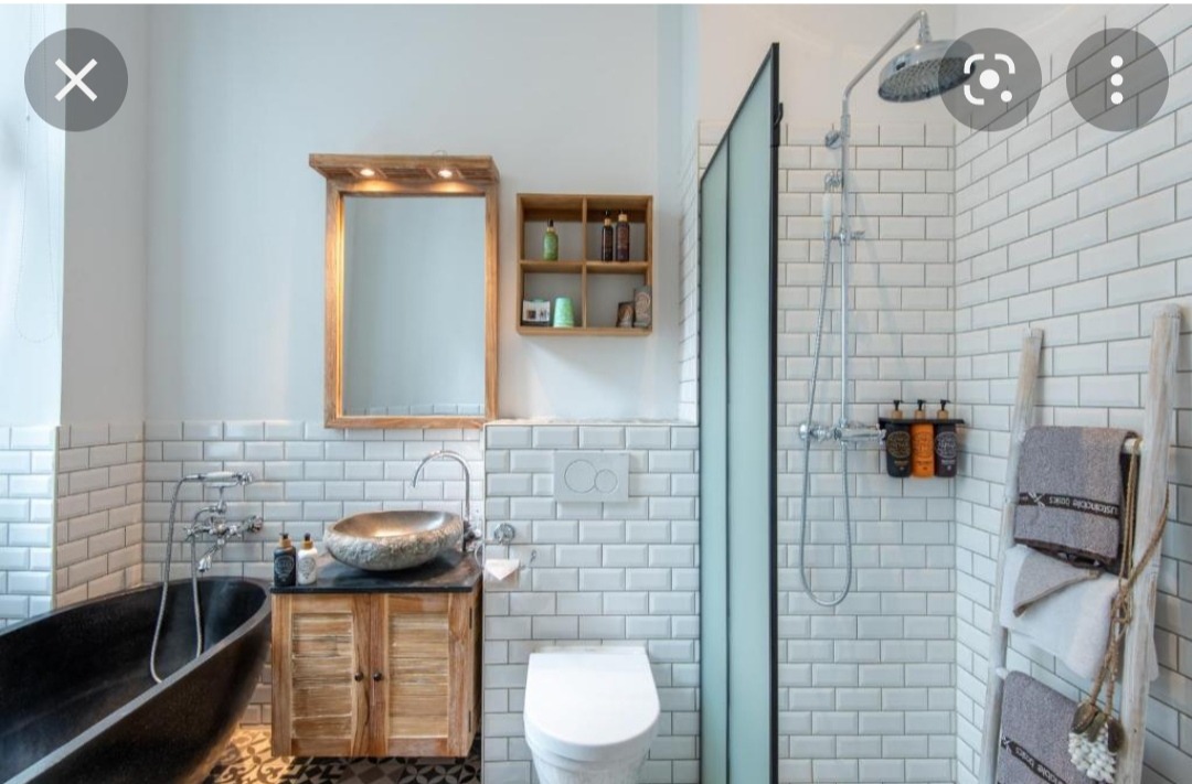 Nowoczesna łazienka z czarną wanną, umywalką z kamienia na drewnianej szafce, białą toaletą, kabiną prysznicową i drabinką na ręczniki, wykończona białymi płytkami imitującymi cegłę.