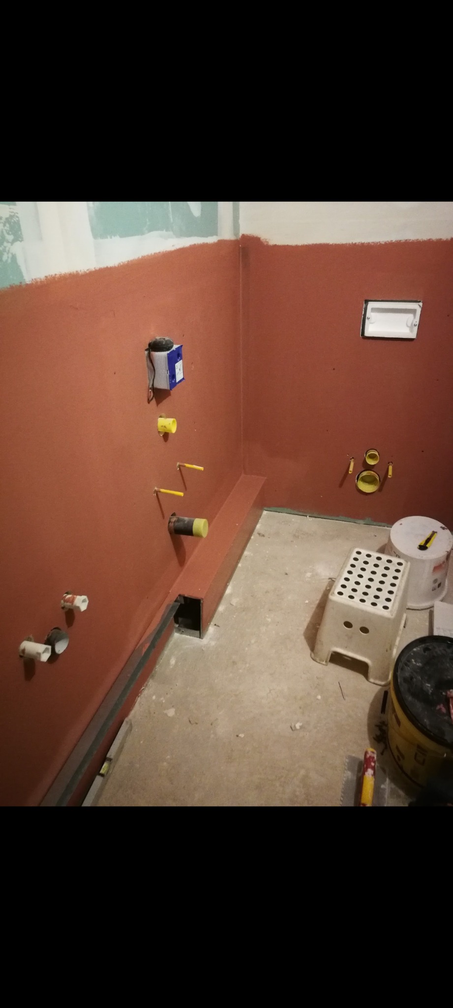 Przygotowanie ścian do wykończenia w łazience: ściany pokryte czerwoną warstwą izolacyjną, widoczne instalacje wodne i elektryczne, oraz narzędzia na podłodze.