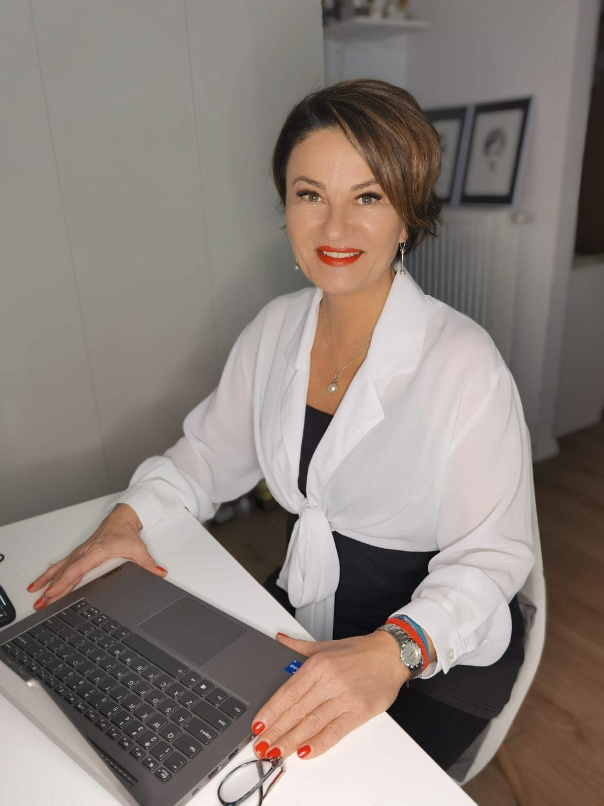 Kobieta w białej koszuli siedzi przy biurku z laptopem, uśmiecha się, ma czerwone usta i paznokcie, na nadgarstku zegarek i dwie bransoletki, na biurku okulary.