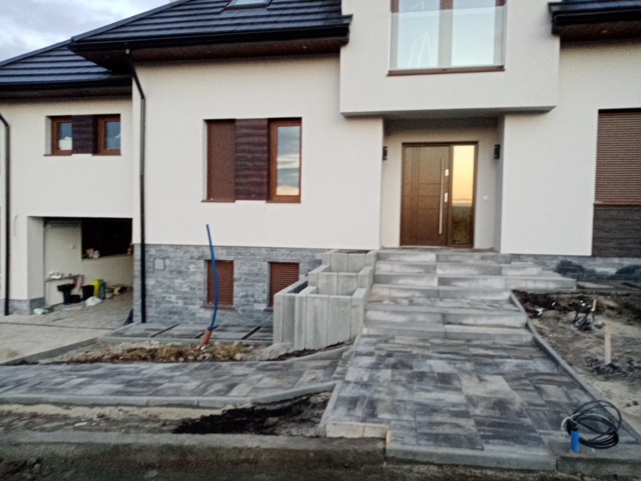 Nowoczesny dom jednorodzinny z szarą kostką brukową ułożoną na podjeździe i schodach wejściowych, widoczne niedokończone prace ziemne i narzędzia, błękitna rura wystająca z ziemi.
