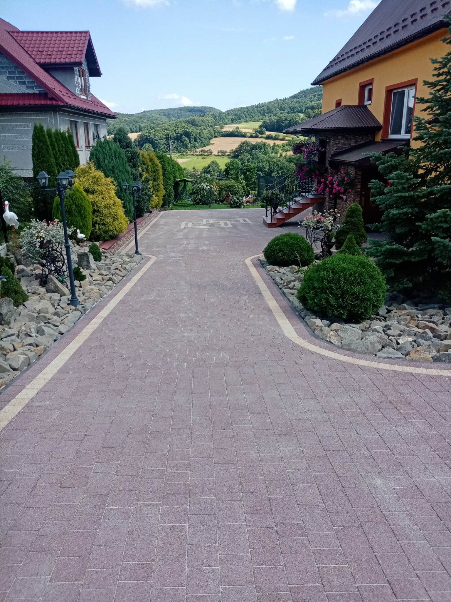 Szeroka, nowo ułożona ścieżka z kostki brukowej prowadząca do dwóch domów, otoczona ozdobnymi kamieniami, roślinami i lampami ogrodowymi, z widokiem na pagórkowaty krajobraz w tle.