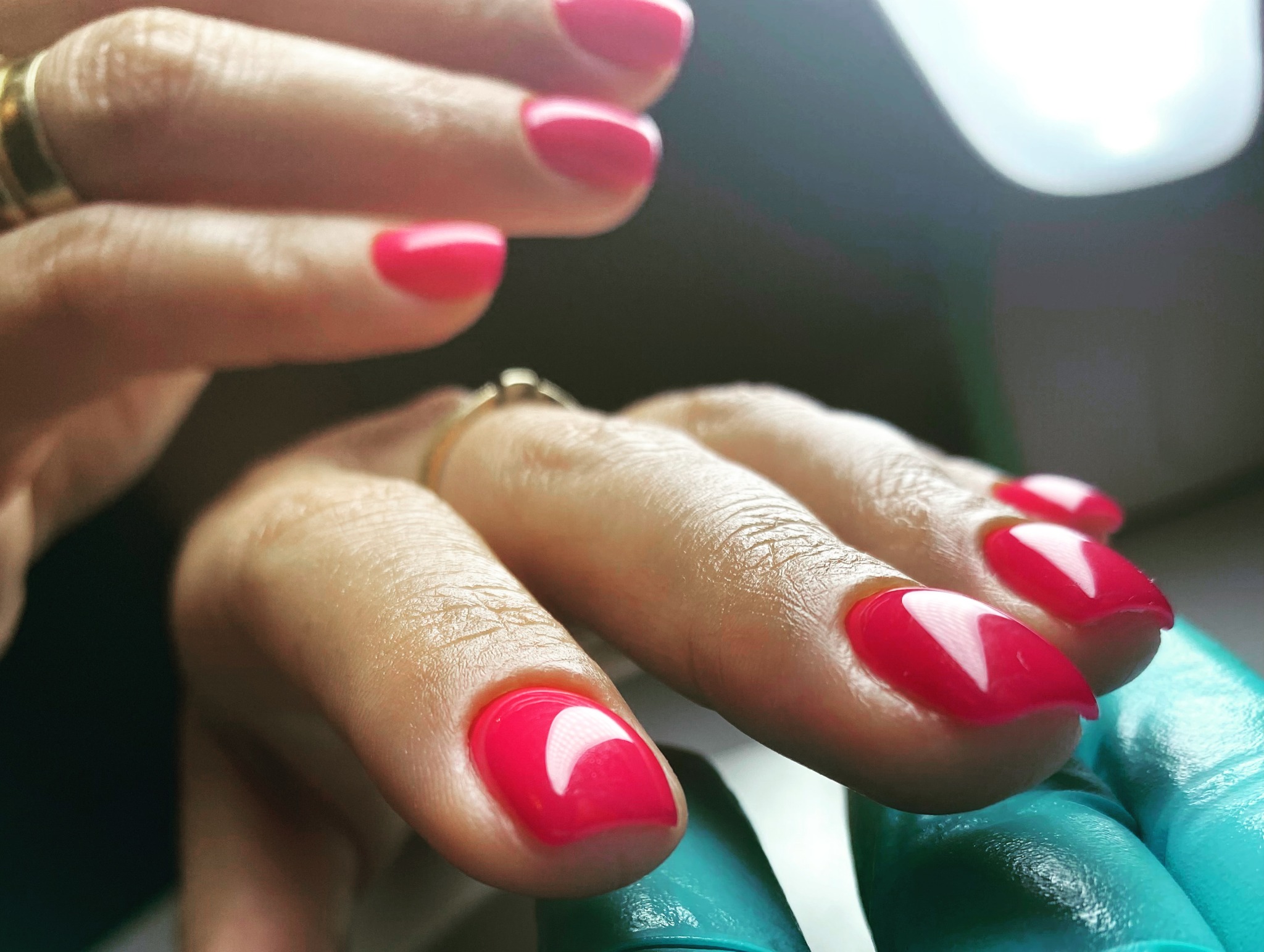Zbliżenie na dłoń z jaskraworóżowym manicurem żelowym o kwadratowym kształcie, z widocznym pierścionkiem na palcu wskazującym i dłonią w turkusowej rękawiczce w tle.