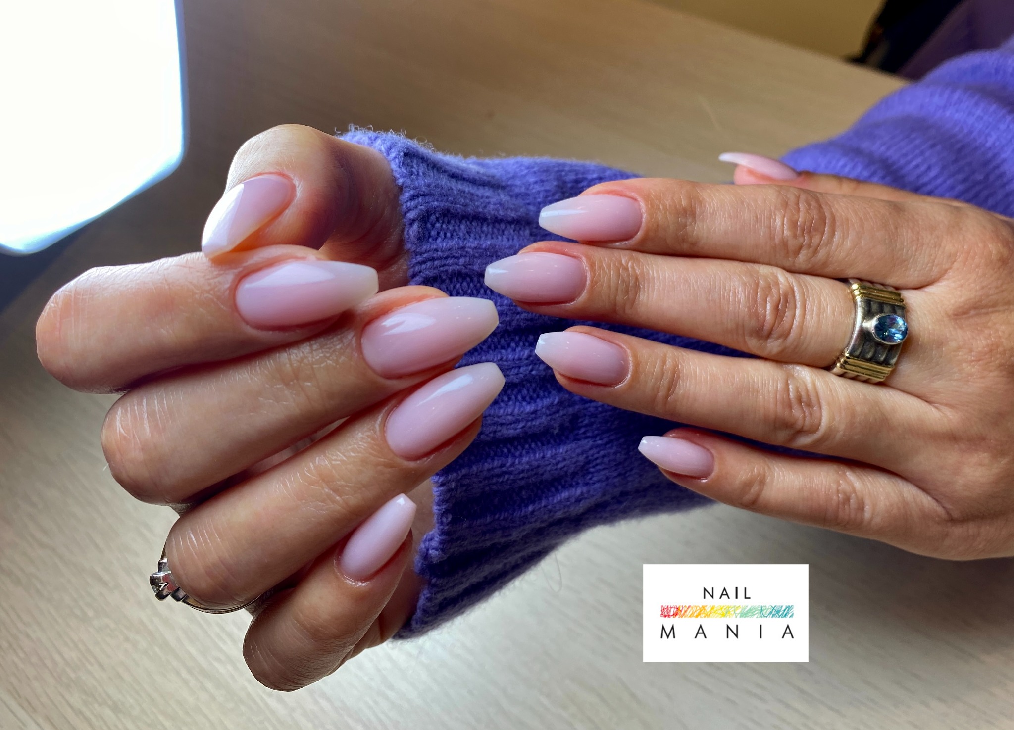 Dłonie z subtelnym, jasnoróżowym manicure hybrydowym w kształcie migdała, pierścionek z niebieskim kamieniem na serdecznym palcu, fioletowy sweter w tle.