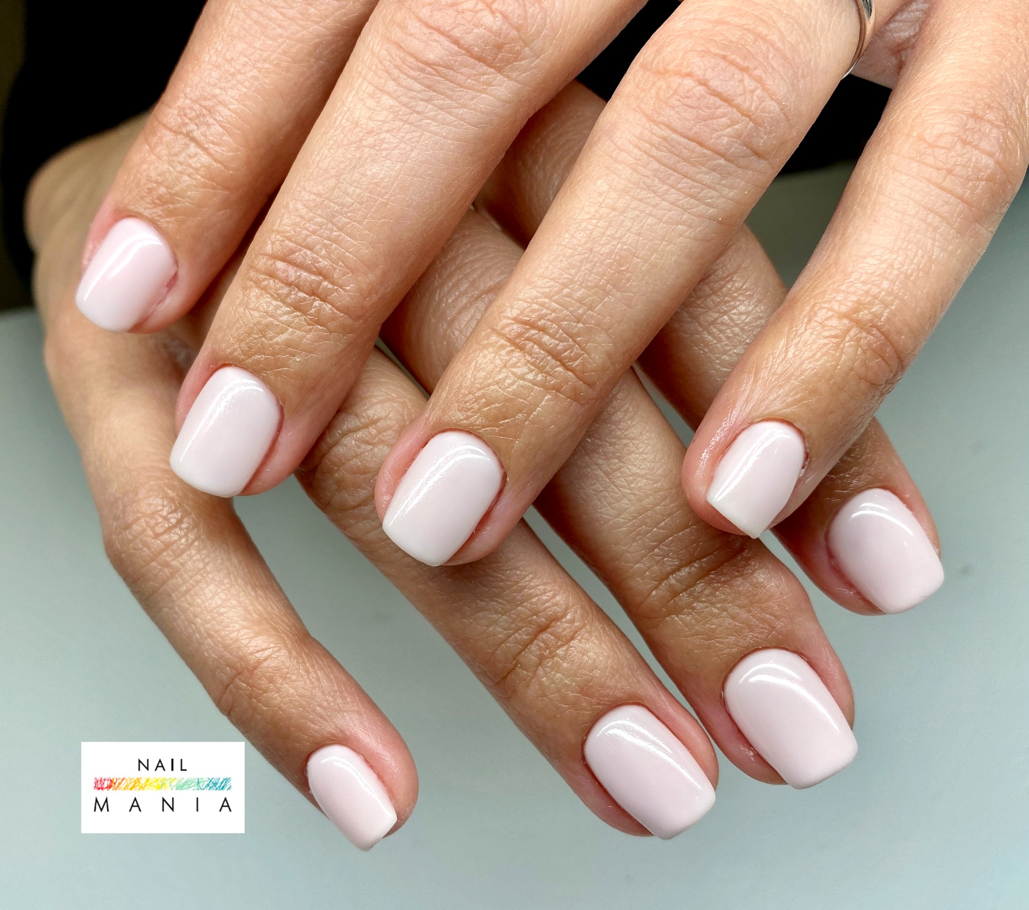 Dłonie ze świeżo pomalowanymi paznokciami w jasnoróżowym odcieniu, kwadratowy kształt, widoczny pierścionek na palcu wskazującym.