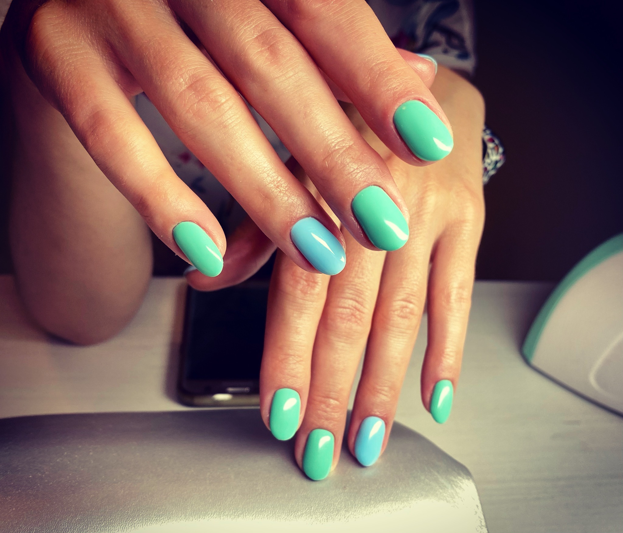 Dłonie z świeżo pomalowanymi paznokciami w odcieniach turkusu i błękitu, na tle lampy UV do utwardzania lakieru hybrydowego i smartfona.