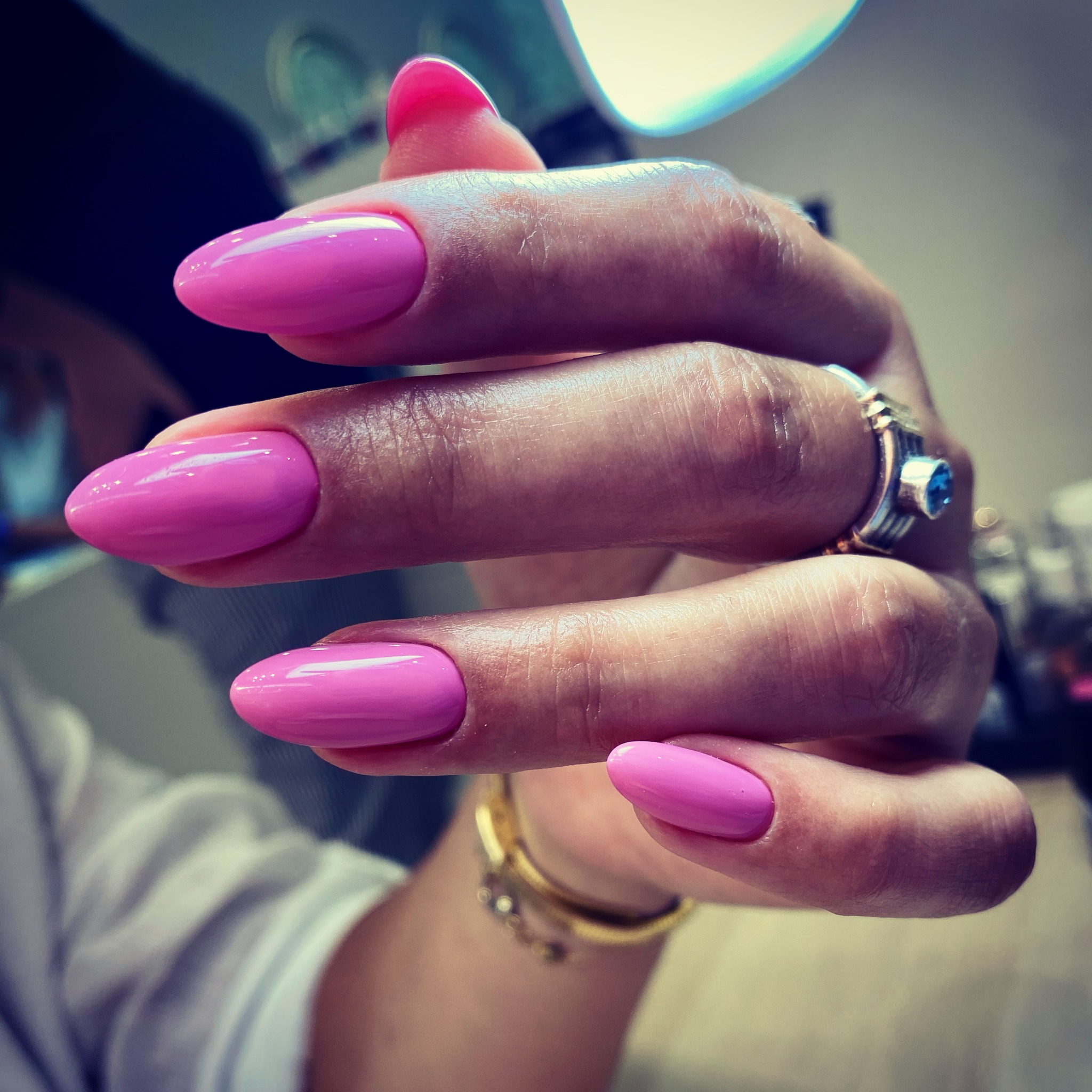 Dłoń z świeżo pomalowanymi, różowymi paznokciami w kształcie migdała, ozdobiona pierścionkiem z niebieskim oczkiem i złotą bransoletką, prezentowana w salonie w Krakowie.