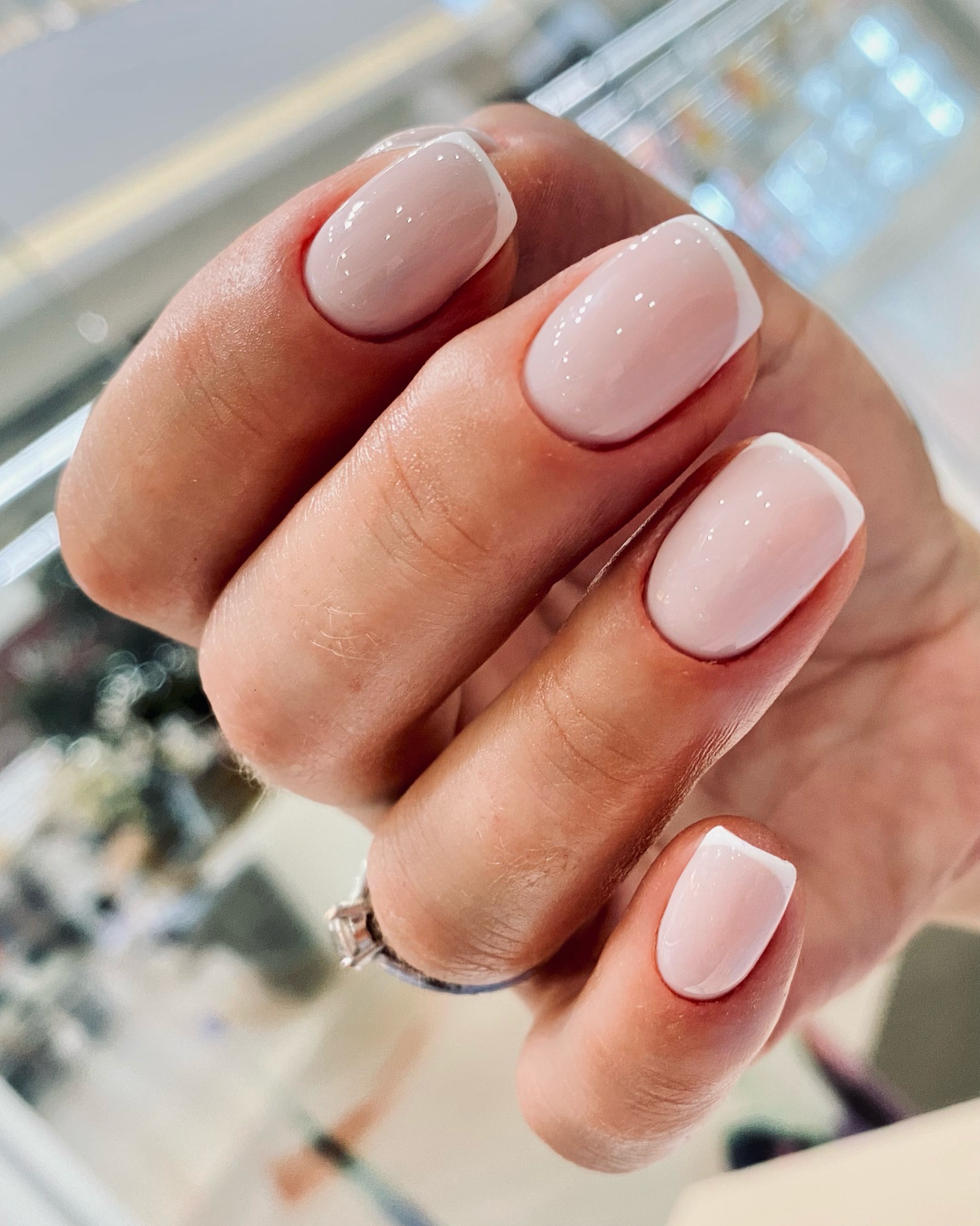 Dłoń z klasycznym, jasnoróżowym manicure francuskim na krótkich, zaokrąglonych paznokciach, ozdobiona pierścionkiem z pojedynczym kamieniem.